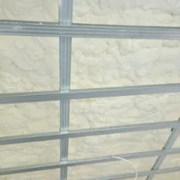 Ocieplenie poddasza pianką PUR, widoczne metalowe profile konstrukcyjne sufitu podwieszanego, biała pianka wypełnia przestrzenie między profilami.