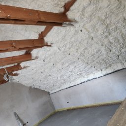 Seven Pur - Poddasze ocieplone białą pianką PUR, widoczne drewniane belki konstrukcyjne, szpilki montażowe, fragment ściany i podłogi w surowym stanie.