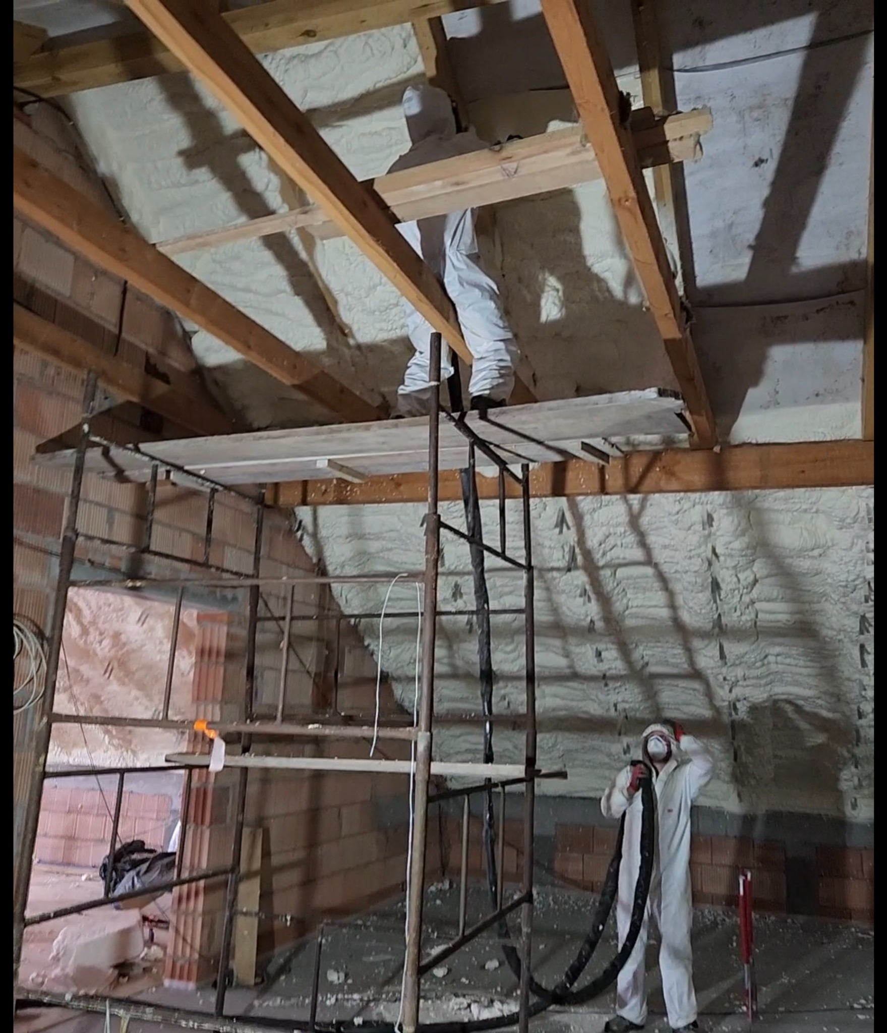Pracownicy w kombinezonach ochronnych aplikują izolację natryskową na poddaszu, widoczne rusztowanie i drewniana konstrukcja dachu.