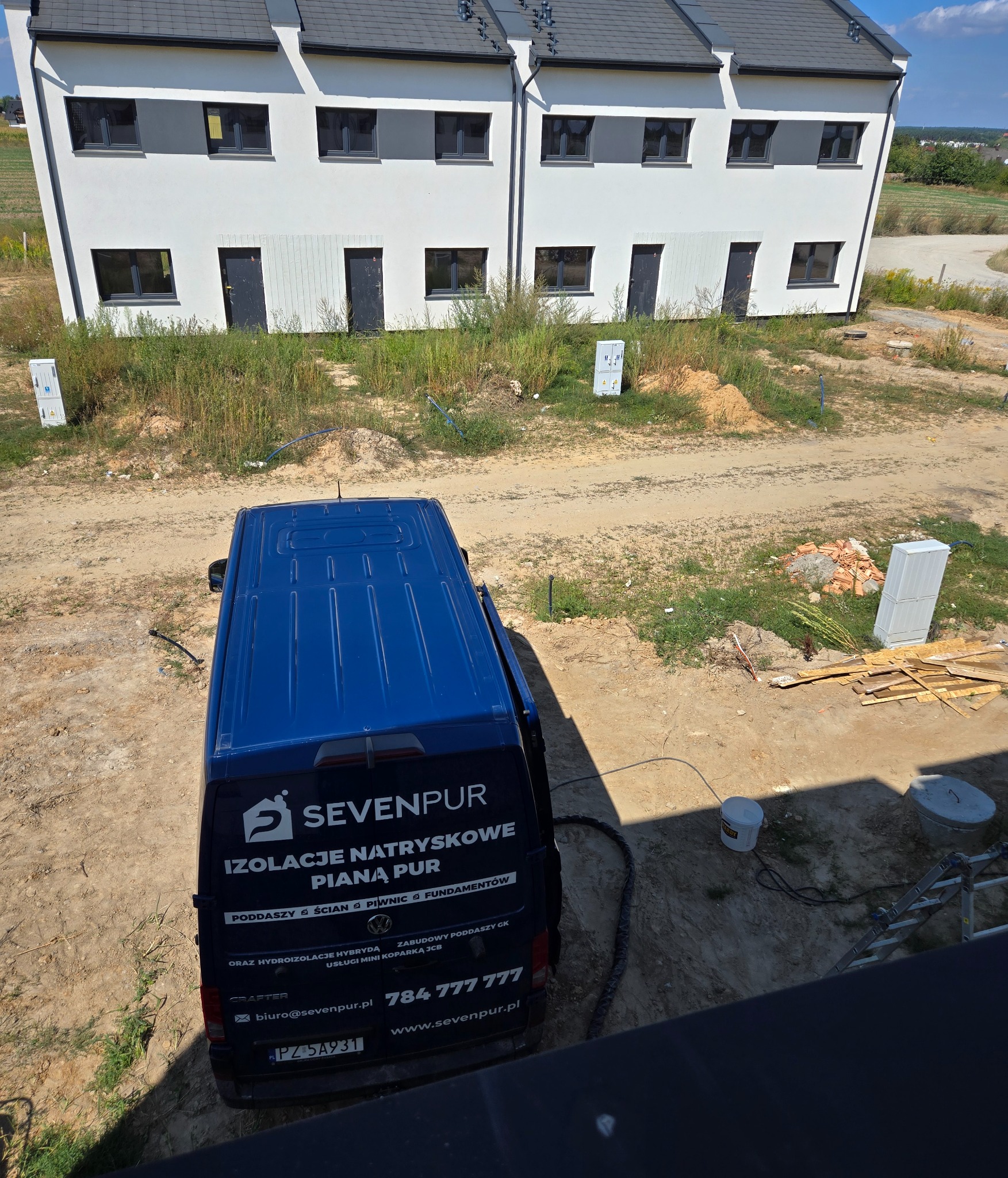 Widok z góry na zaparkowany niebieski samochód dostawczy z logo firmy Sevenpur, świadczącej usługi izolacji natryskowej pianą PUR, na tle nowo wybudowanych, surowych domów jednorodzinnych...