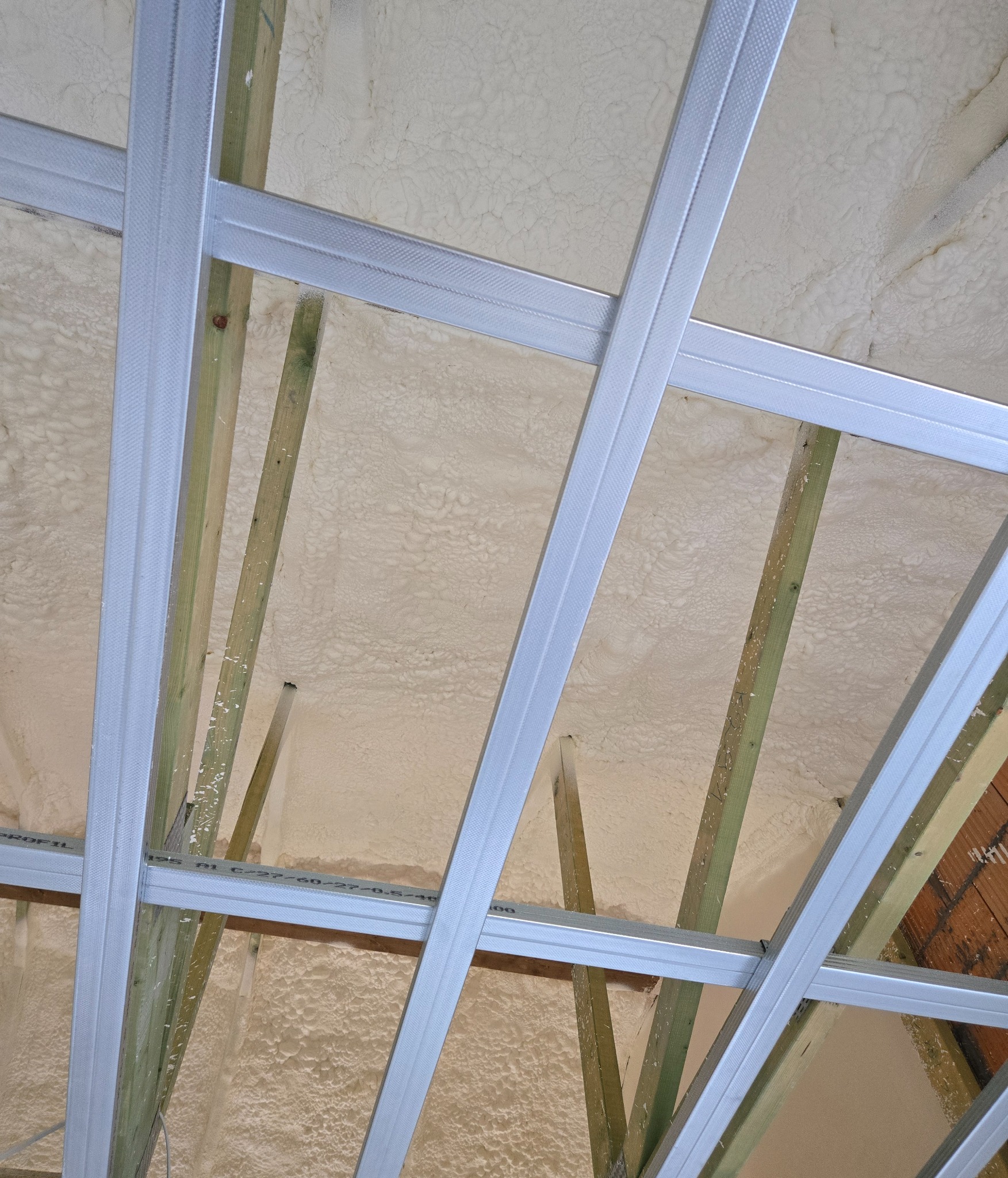 Klatka sufitowa z profili metalowych i drewnianych belek wypełniona jasną, nierówną izolacją natryskową.