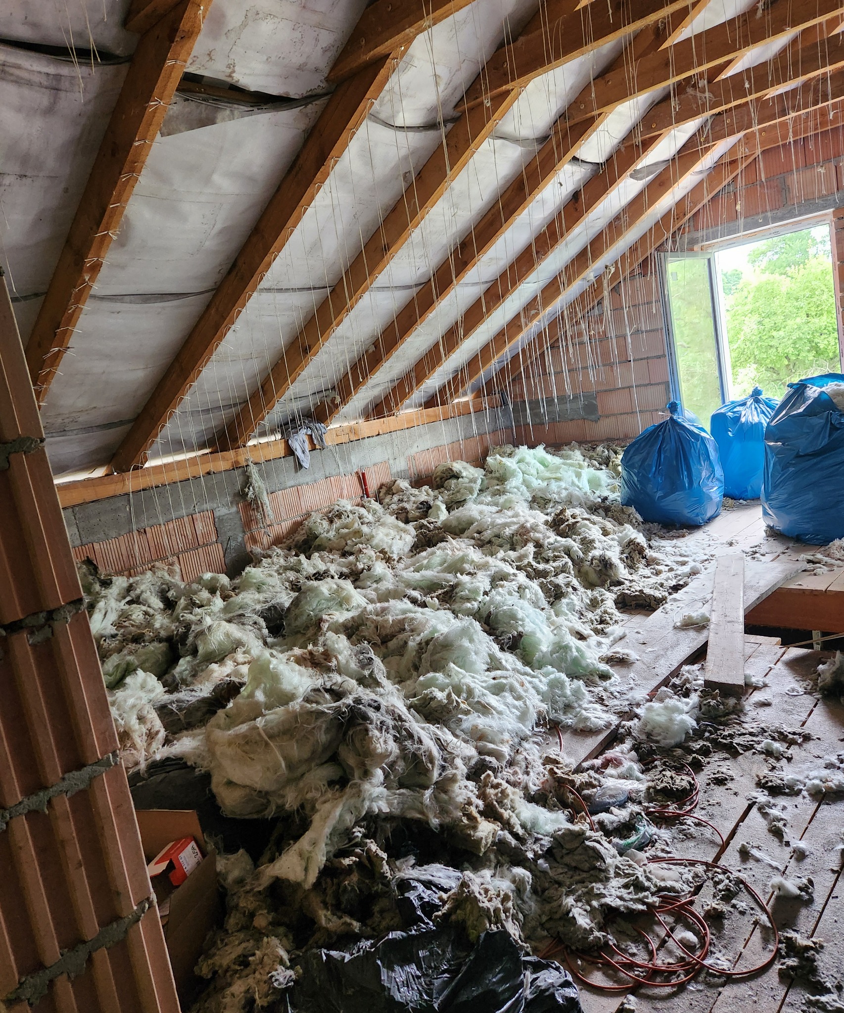 Poddasze w trakcie prac izolacyjnych: widoczne drewniane krokwie z mocowaniami, warstwa wstępnej izolacji, rozsypane włókna izolacyjne oraz niebieskie worki z materiałem. Ujęcie z dołu, widoczne...