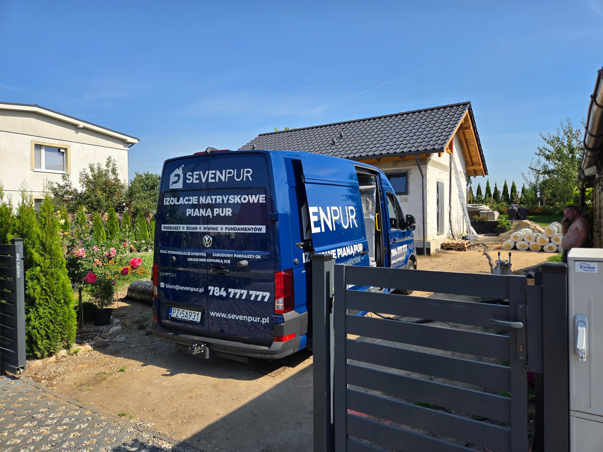 Niebieski samochód dostawczy z reklamą firmy izolacyjnej na tle domu w trakcie budowy i materiałów izolacyjnych.