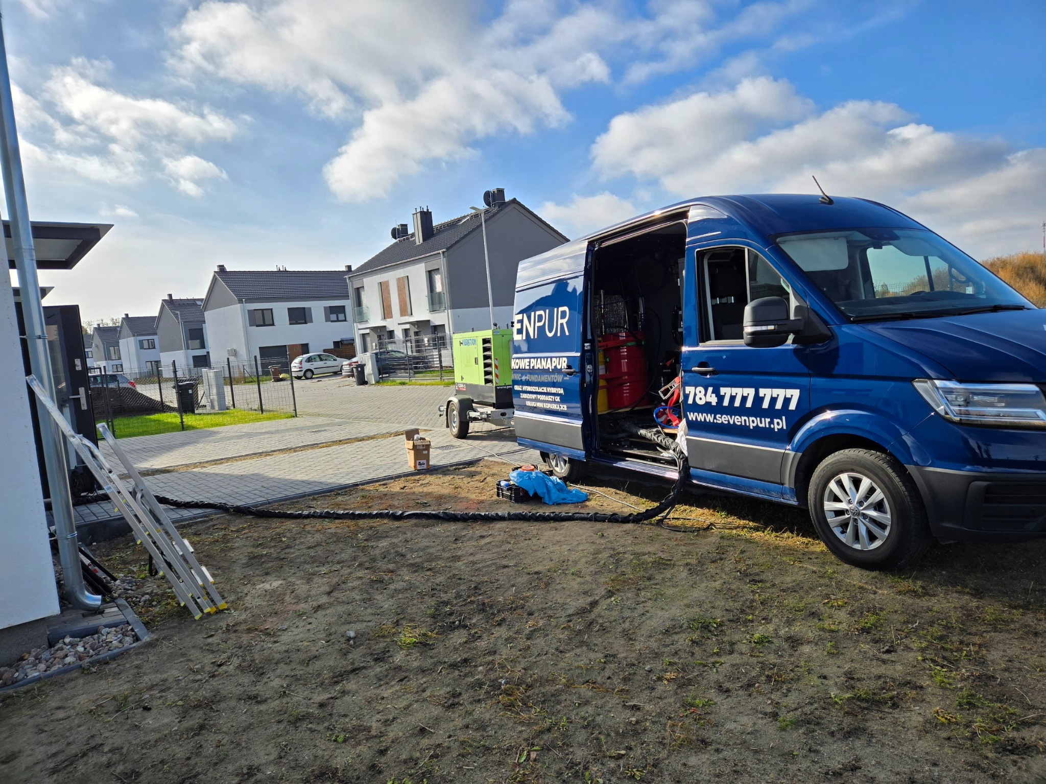 Niebieski van z otwartymi drzwiami, zaparkowany obok domu, z widocznym sprzętem do natrysku pianki i generatorem na przyczepie, przygotowany do pracy.