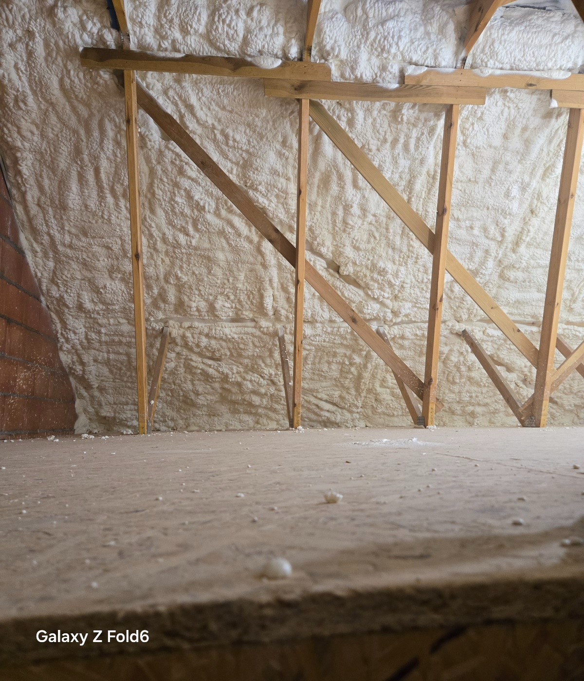 Poddasze z drewnianą konstrukcją i nałożoną białą pianą izolacyjną, widok z dołu na surową podłogę z resztkami piany.