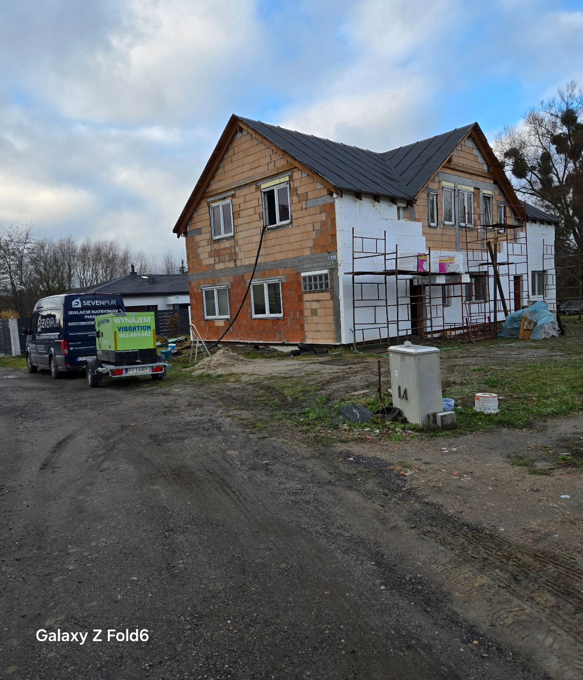 Dom w trakcie ocieplania, widoczne rusztowania, częściowo ocieplona ściana, zaparkowany van z napisem 'Izolacje natryskowe pianą', obok przyczepka z agregatem natryskowym.