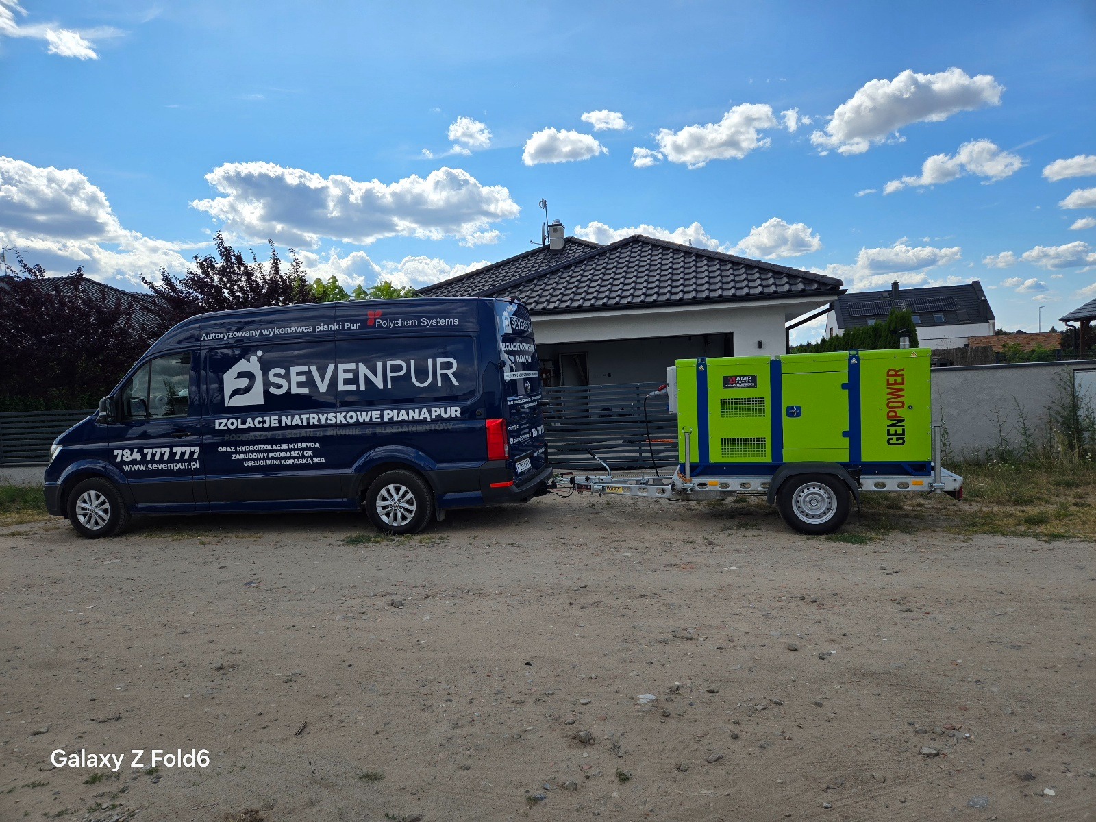 Granatowy van Sevenpur z przyczepą i agregatem prądotwórczym na tle domu jednorodzinnego i błękitnego nieba. Usługi izolacji natryskowej pianą PUR.