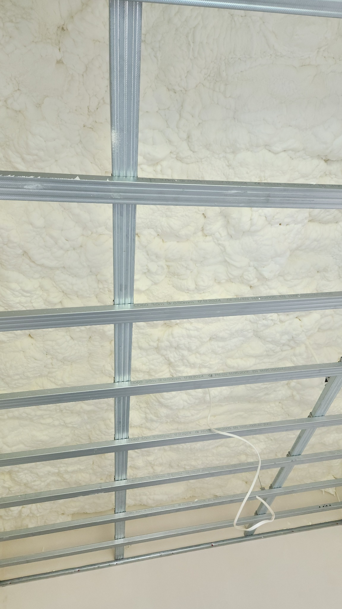 Ocieplenie poddasza pianką PUR, widoczne metalowe profile konstrukcyjne sufitu podwieszanego, biała pianka wypełnia przestrzenie między profilami.