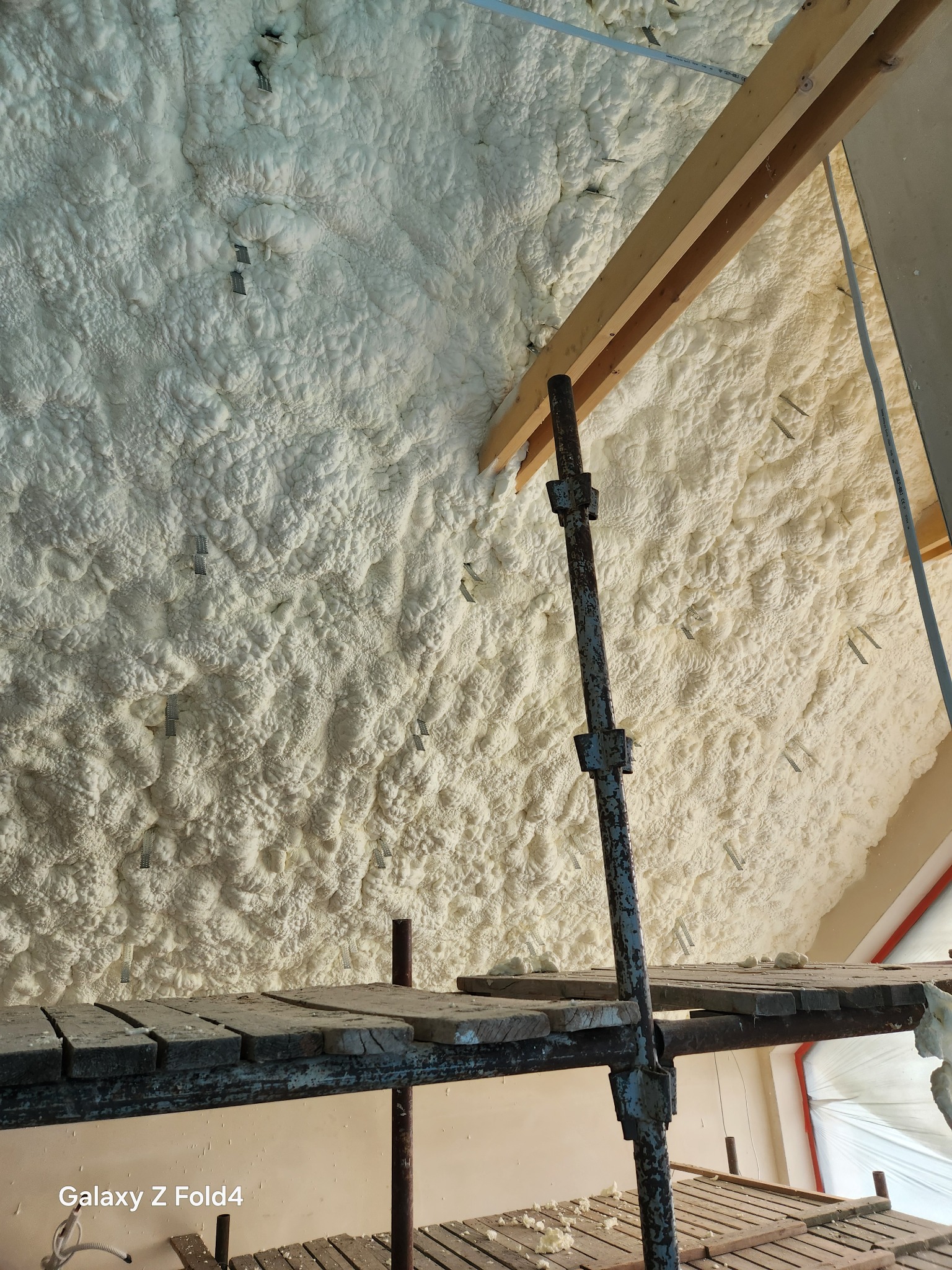 Poddasze ocieplone pianką PUR, widoczne metalowe mocowania, drewniana konstrukcja i rusztowanie. Aplikacja pianki na ukośnej powierzchni.