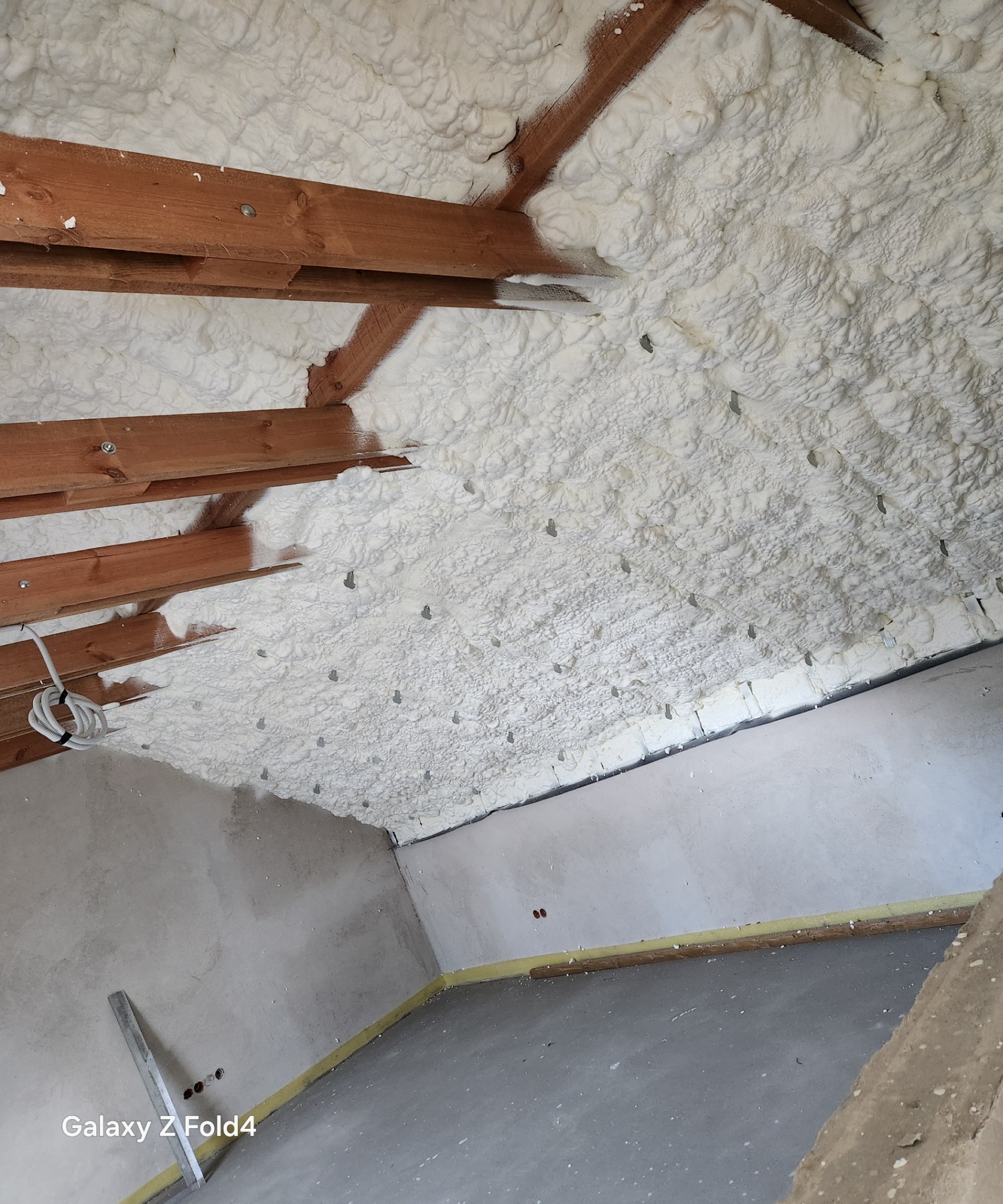 Poddasze ocieplone białą pianką PUR, widoczne drewniane belki konstrukcyjne, szpilki montażowe, fragment ściany i podłogi w surowym stanie.