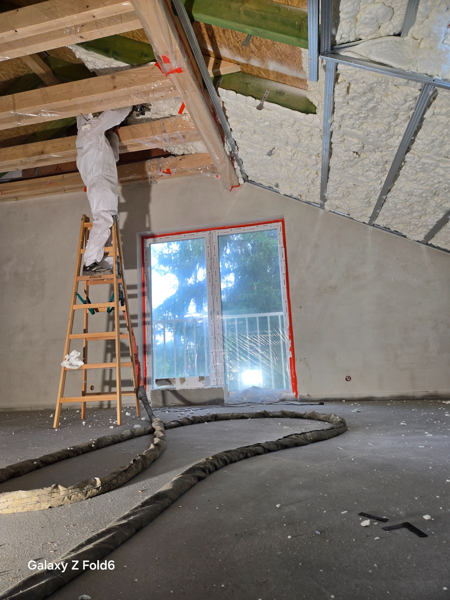 Aplikacja pianki PUR na poddaszu w Kórniku. Pracownik w kombinezonie na drabinie. Widoczna warstwa izolacyjna i przewód doprowadzający piankę na podłogę.