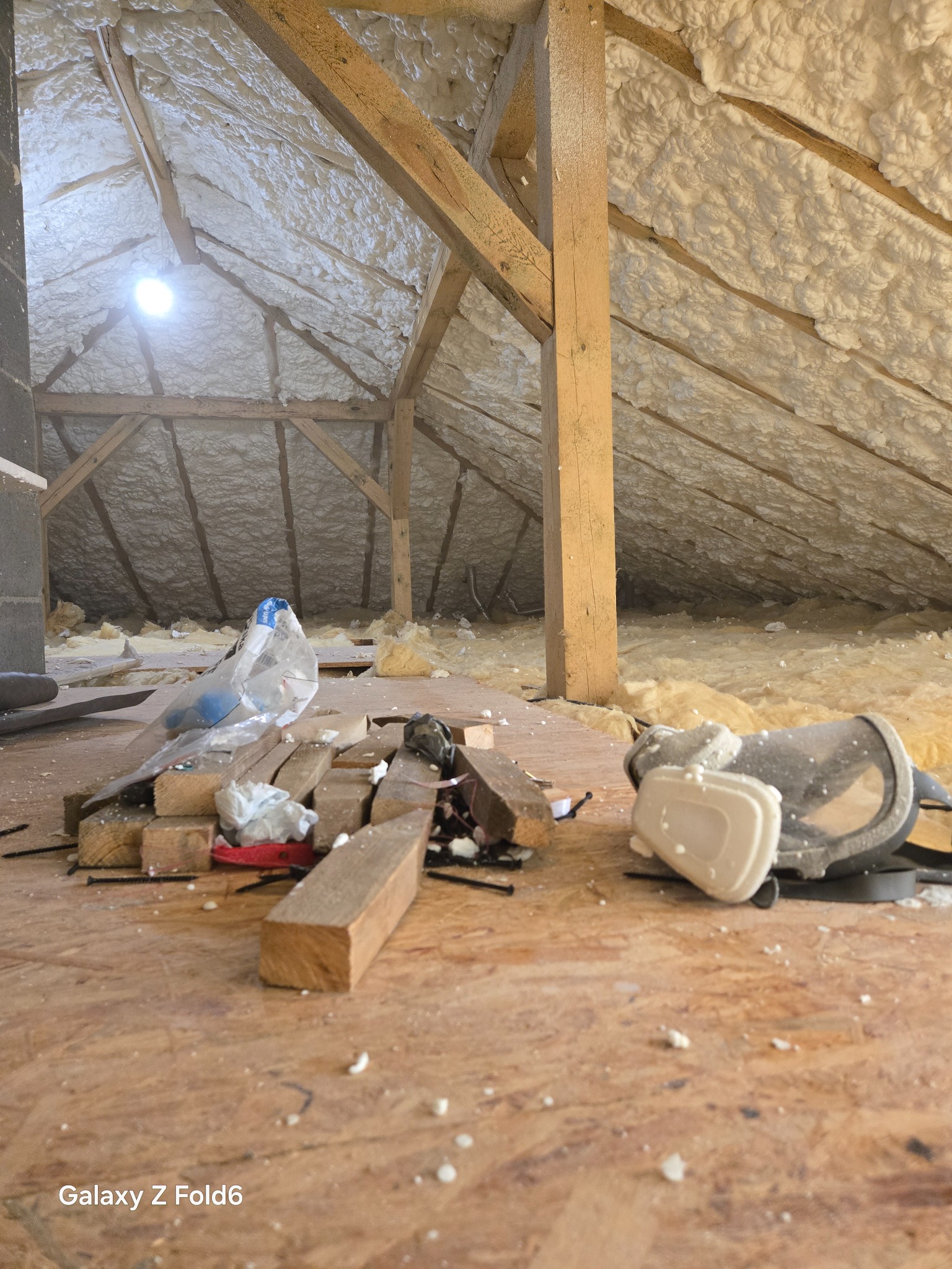 Poddasze ocieplone pianką PUR, z widocznymi drewnianymi belkami konstrukcyjnymi. Na podłodze leżą resztki materiałów budowlanych i maska ochronna.