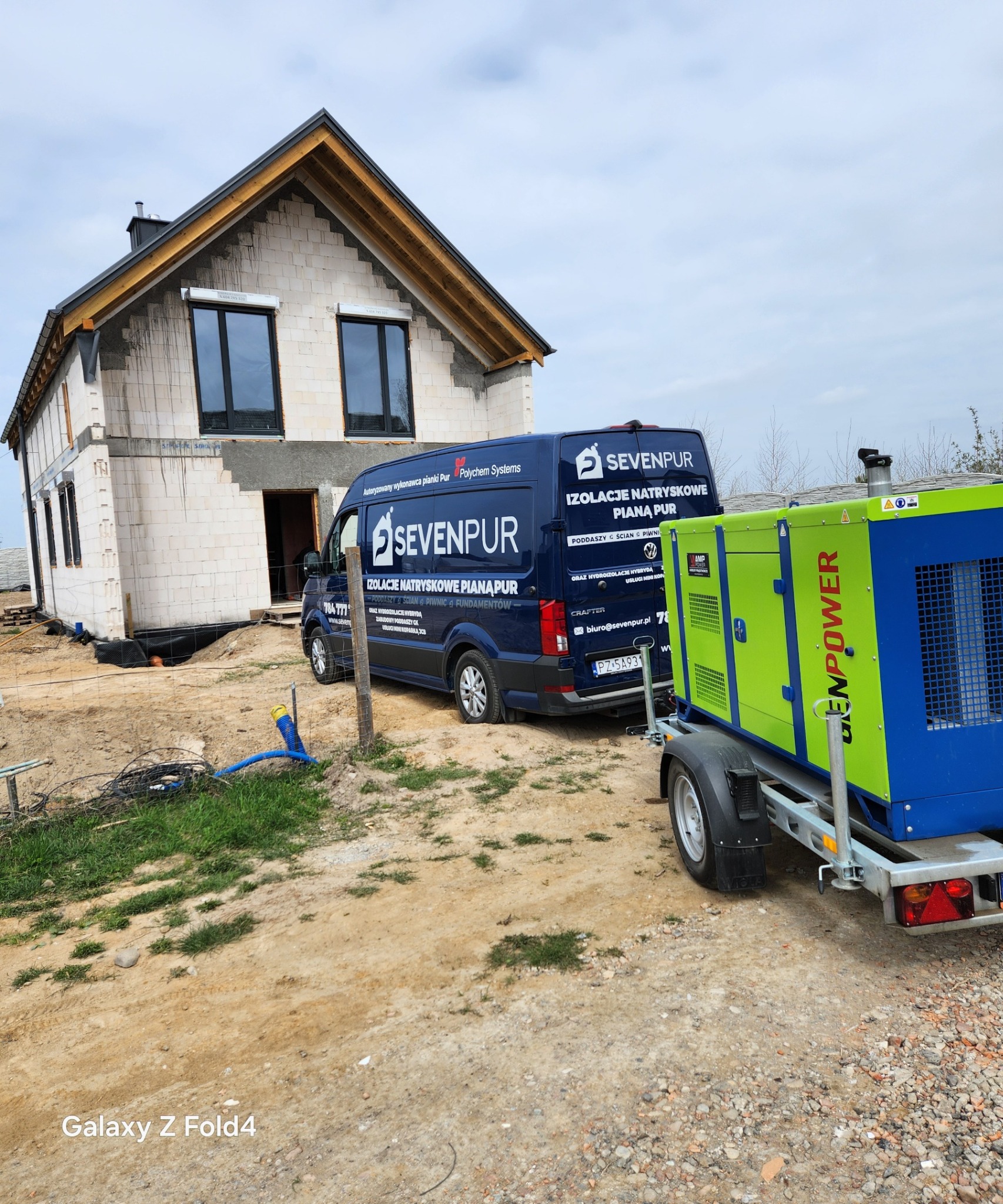 Dom w trakcie ocieplania pianką PUR. Na posesji stoi bus firmy Sevenpur i agregat prądotwórczy. Widoczne niezabezpieczone fundamenty i ściany z pustaków.