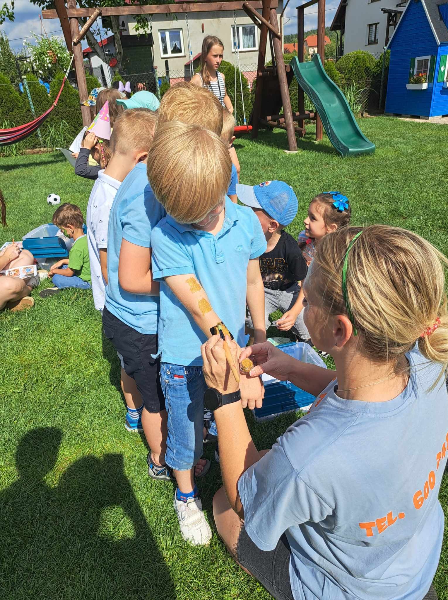 Malowanie brokatowych tatuaży na ramieniu dziecka podczas imprezy plenerowej, w tle grupa dzieci, zjeżdżalnia i huśtawka.