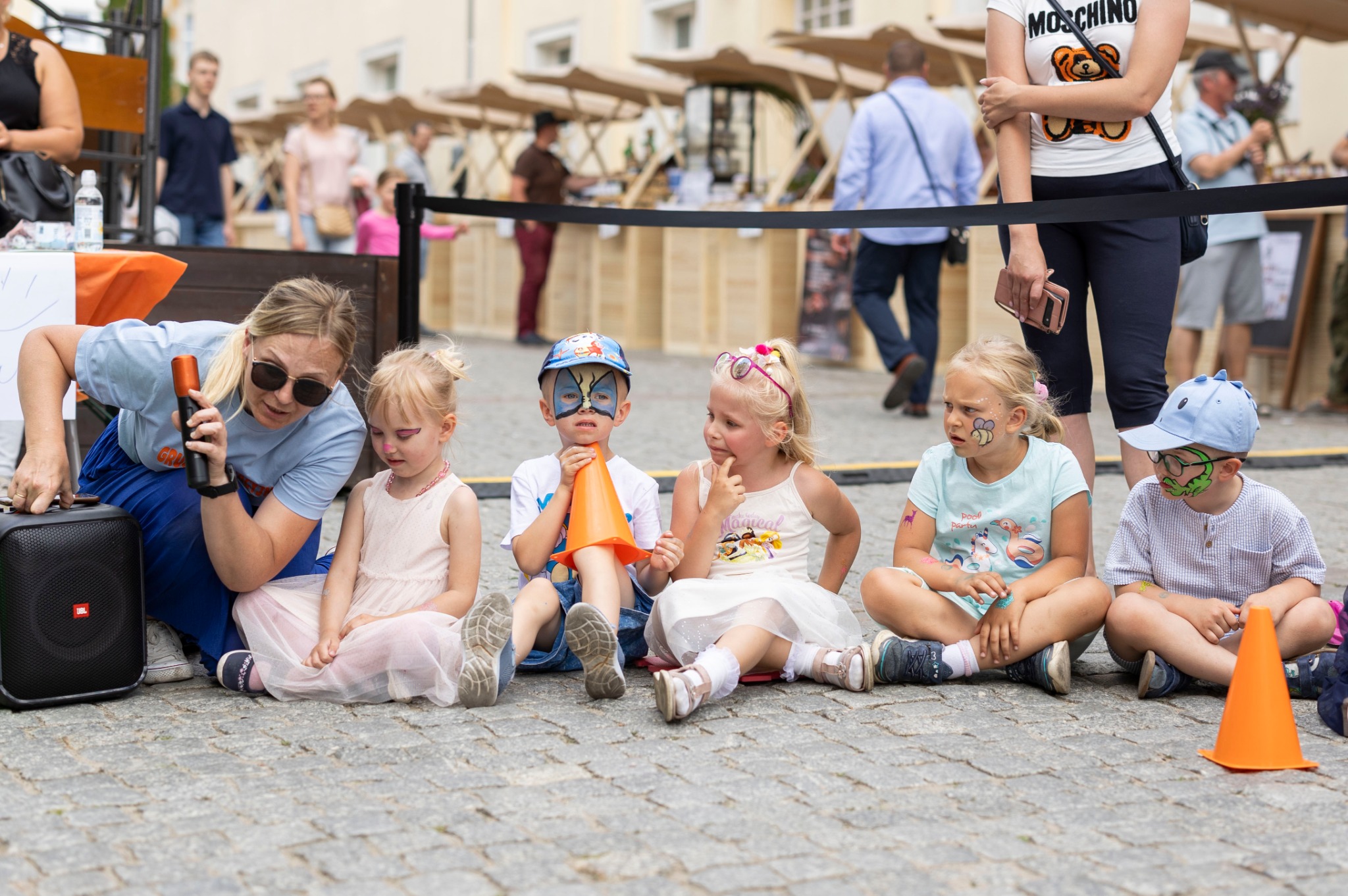 Animator z mikrofonem prowadzi zabawy dla dzieci z pomalowanymi twarzami, siedzących na brukowanej powierzchni podczas wydarzenia plenerowego z drewnianymi stoiskami w tle.