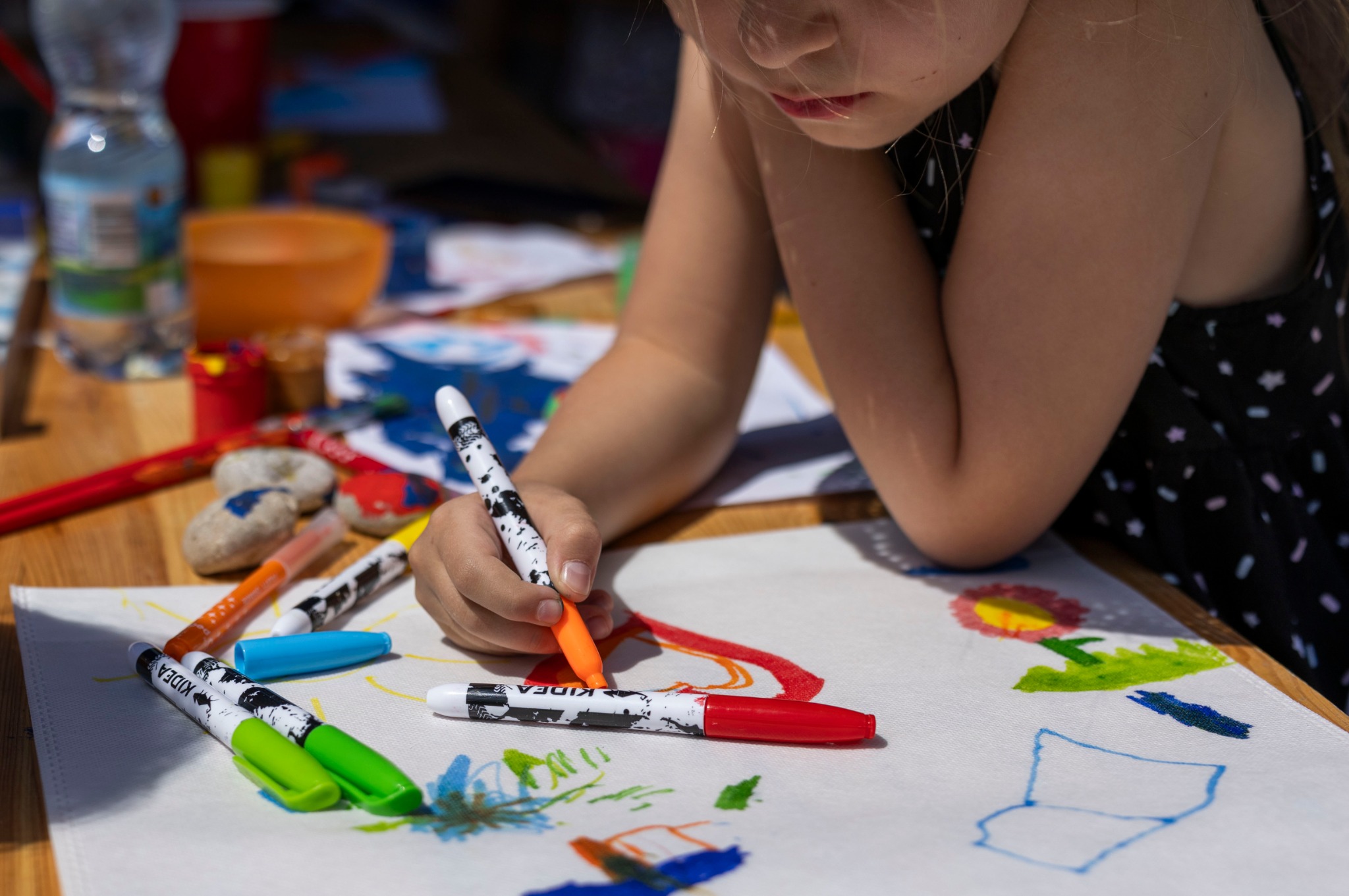 Dziewczynka rysuje pomarańczowym flamastrem na białym płótnie, otoczona kolorowymi pisakami i kamieniami pomalowanymi farbami.