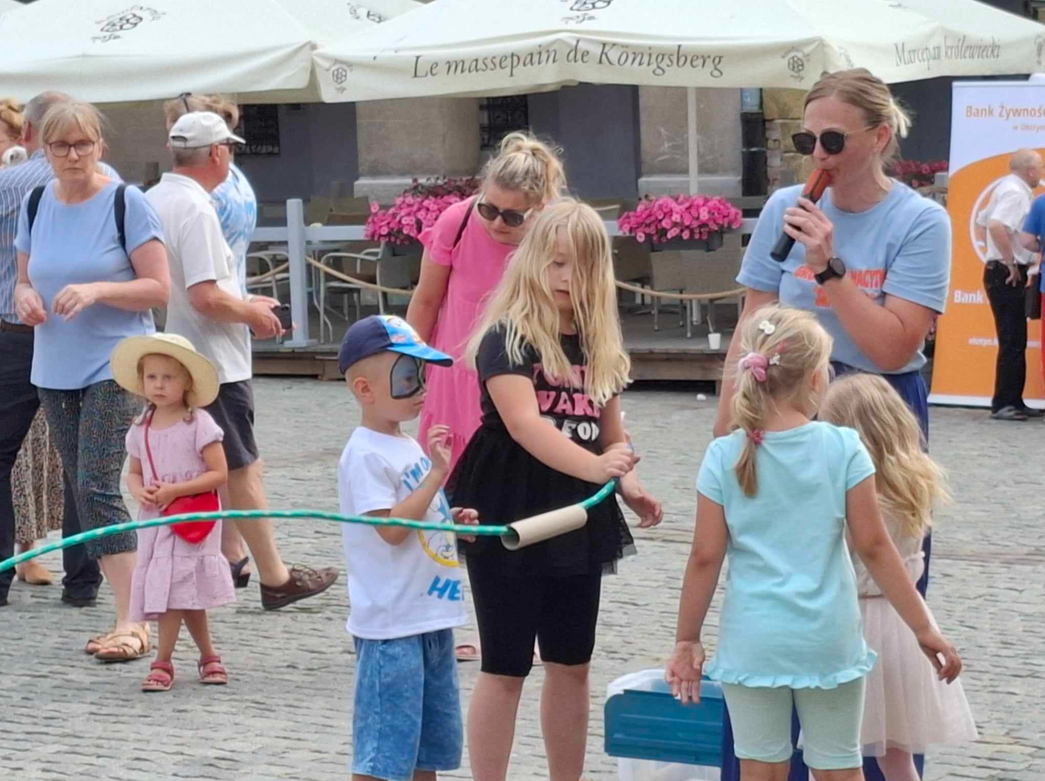 Animator prowadzi zabawy z dziećmi na placu miejskim, wykorzystując rurkę po papierze toaletowym i sznur do przeciągania, w tle parasole kawiarniane.