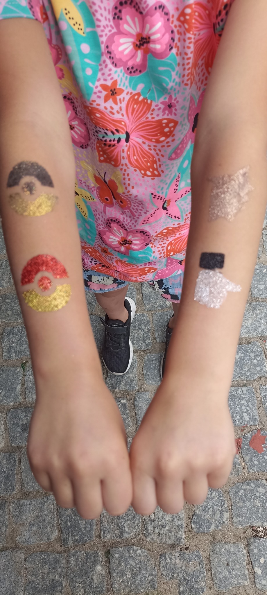 Dziecko prezentuje brokatowe tatuaże na obu ramionach: dwa okrągłe wzory imitujące Pokeballe w kolorach czarno-złotym i czerwono-złotym na lewym ramieniu oraz abstrakcyjne, błyszczące wzory...