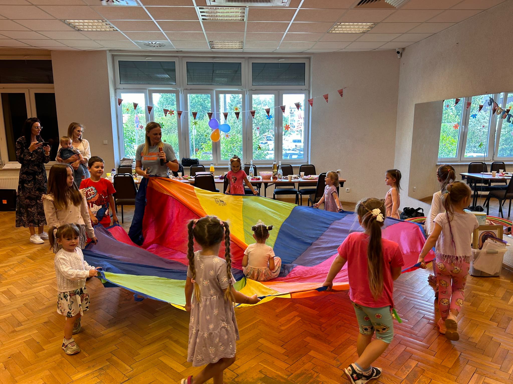 Animator prowadzi zabawę z kolorową chustą animacyjną dla grupy dzieci w sali z parkietem i oknami.