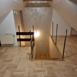 Wykończone poddasze z ułożoną parkietową podłogą w jodełkę, widoczny fragment schodów na niższy poziom z prowizoryczną barierką i oświetleniem.