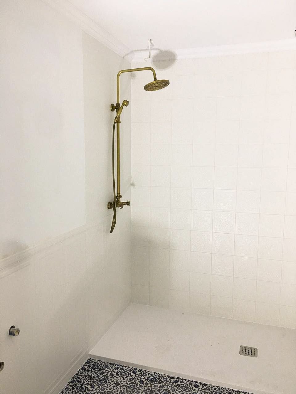 Wyremontowana łazienka z białą kwadratową glazurą, złotą baterią prysznicową i podłogą wykończoną płytkami w czarno-białe wzory.