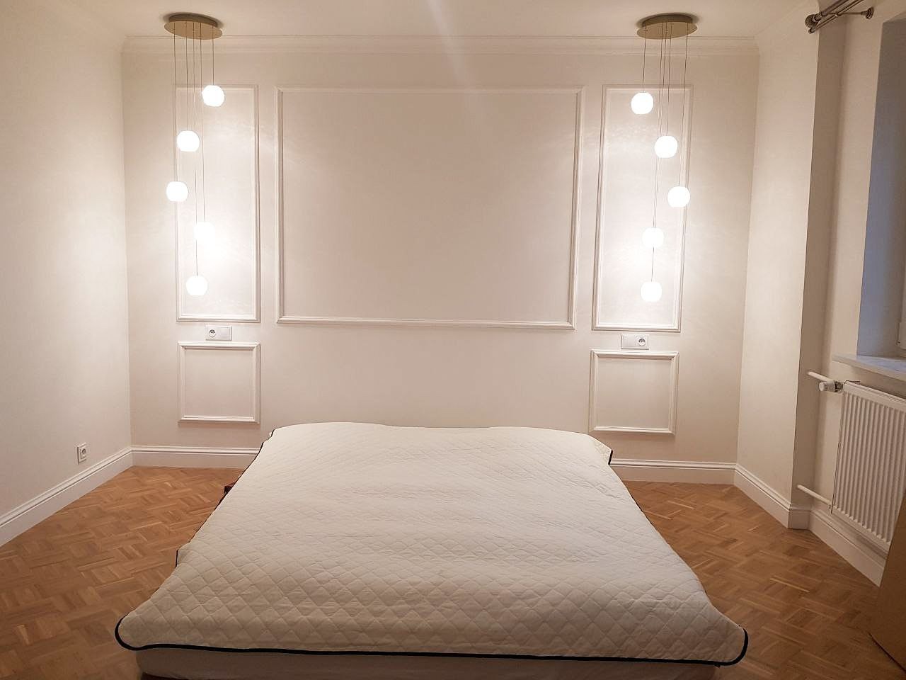 Elegancka sypialnia z białymi ścianami, listwami dekoracyjnymi, parkietem w jodełkę i nowoczesnymi lampami wiszącymi z kulistymi kloszami po obu stronach łóżka.