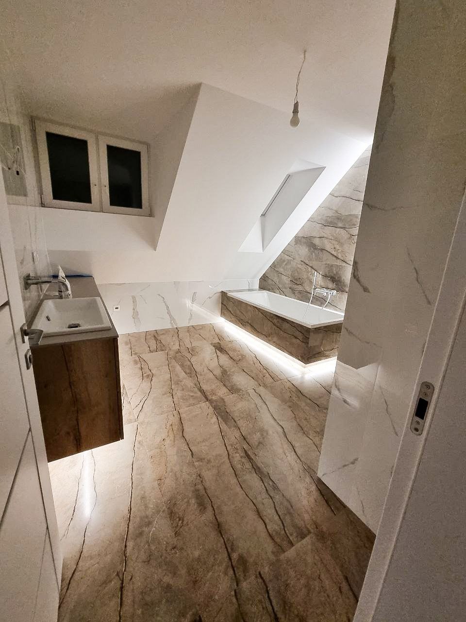 Nowoczesna łazienka na poddaszu z marmurową podłogą i ścianami, wolnostojąca wanna z podświetleniem LED, drewniana szafka pod umywalką i okno dachowe.