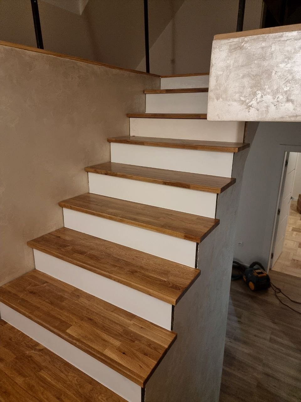 Schody drewniane z białymi podstopnicami, wykończone ścianą o fakturze betonu, widoczny odkurzacz w tle.