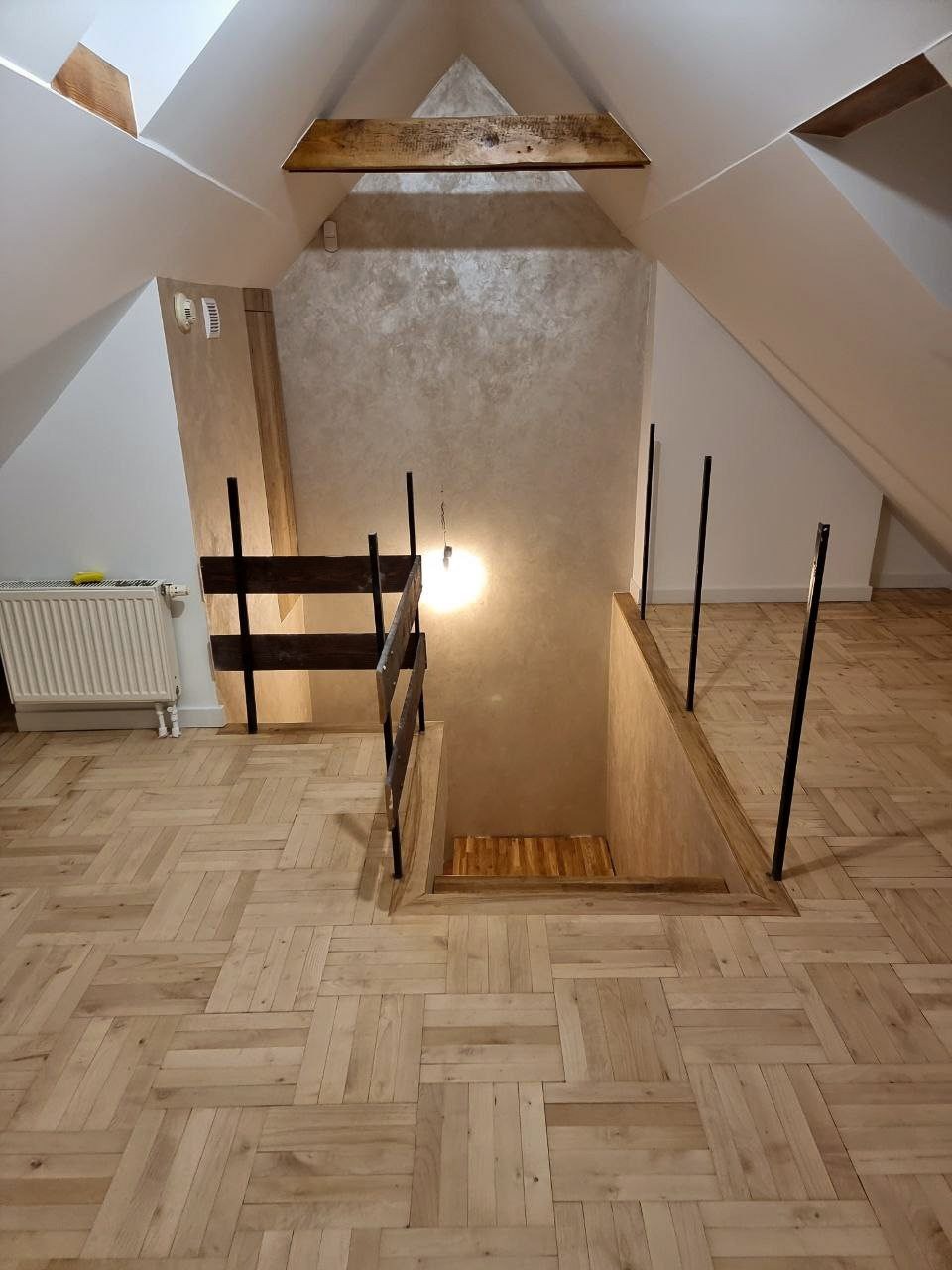 Wykończone poddasze z ułożoną parkietową podłogą w jodełkę, widoczny fragment schodów na niższy poziom z prowizoryczną barierką i oświetleniem.