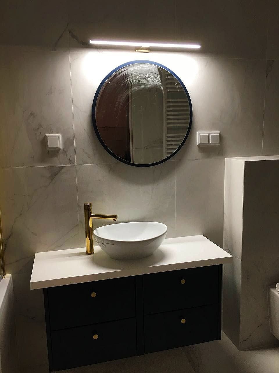 Nowoczesna łazienka z okrągłym lustrem w niebieskiej ramie, podświetlanym ledowym paskiem, białą umywalką nablatową z złotą baterią oraz czarną szafką z białym blatem i złotymi uchwytami na tle...