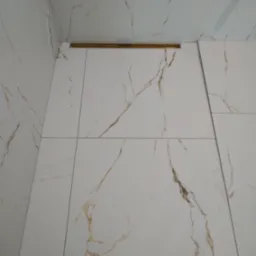 Białe płytki imitujące marmur z delikatnymi złotymi żyłkami, ułożone na ścianach i suficie łazienki, z widoczną złotą listwą dekoracyjną i wystającymi kablami oświetleniowymi.