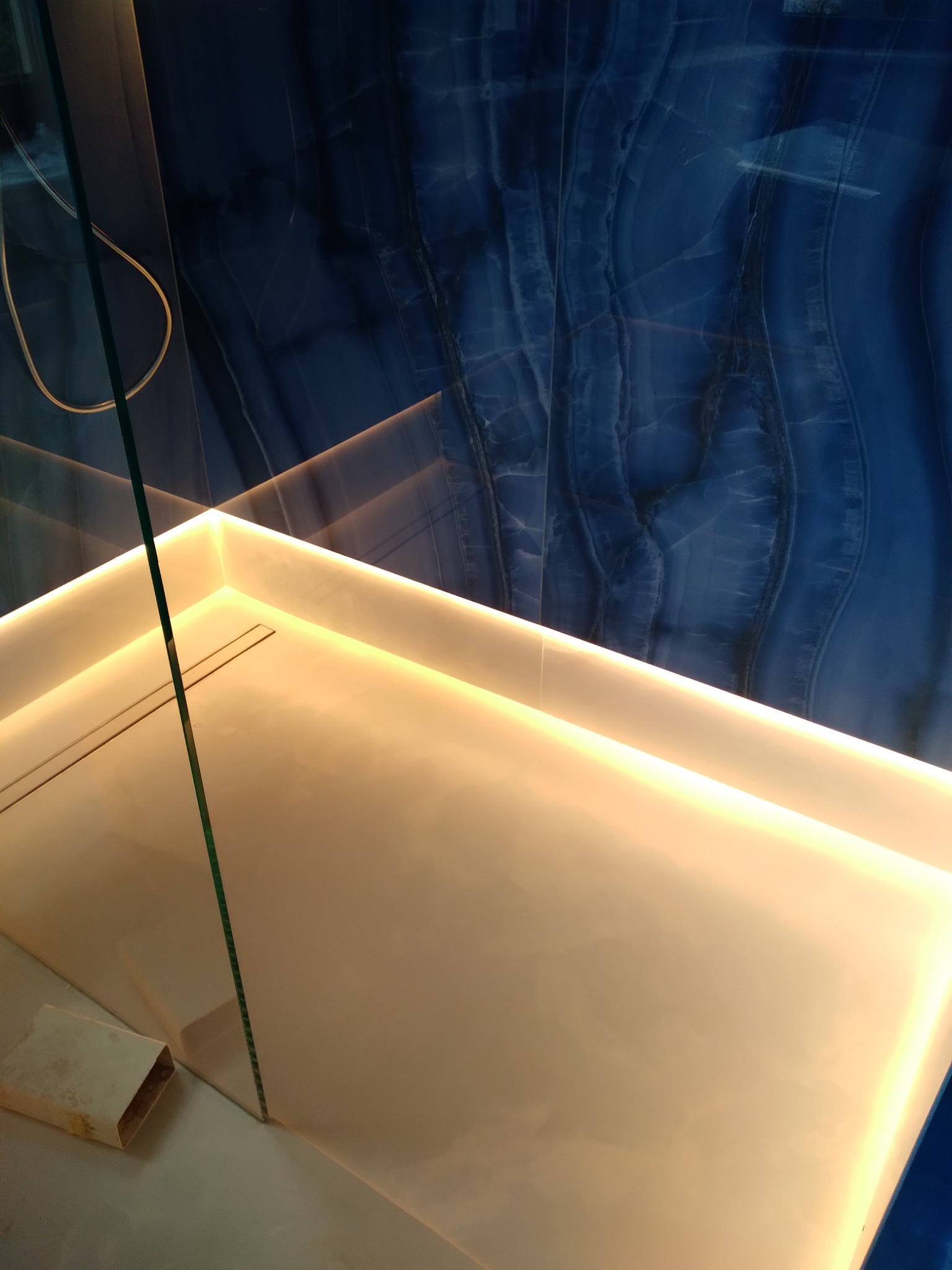 Nowoczesny brodzik z podświetleniem LED i szklaną ścianką kabiny prysznicowej na tle ściany z płytek imitujących niebieski onyks.