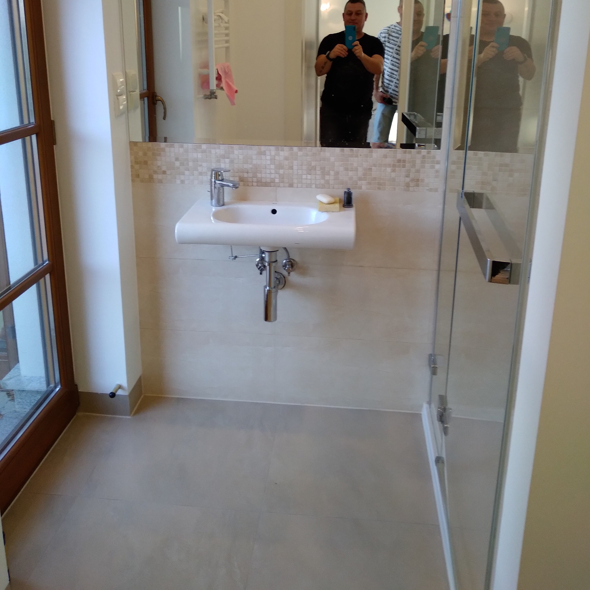 Nowoczesna umywalka z baterią i mydłem na blacie, wykończona mozaiką, widoczna w łazience z kabiną prysznicową i oknem, z odbiciem fotografa w lustrze.