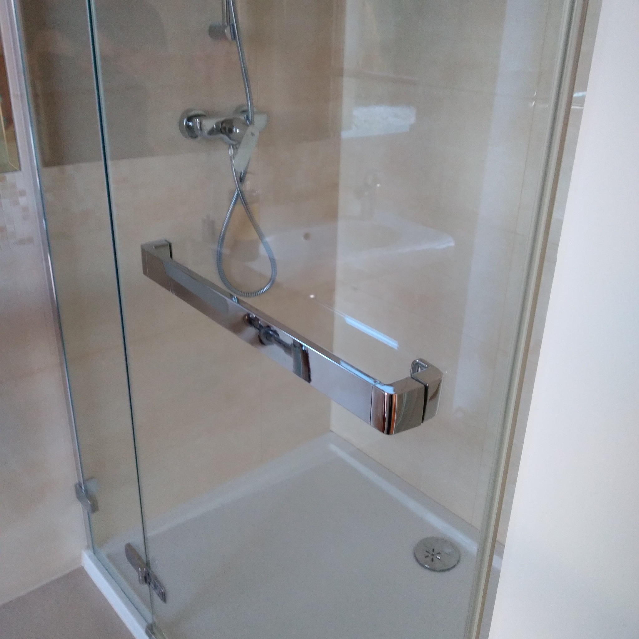 Szklana kabina prysznicowa z chromowanym uchwytem, widoczny brodzik i bateria natryskowa na tle beżowych płytek.