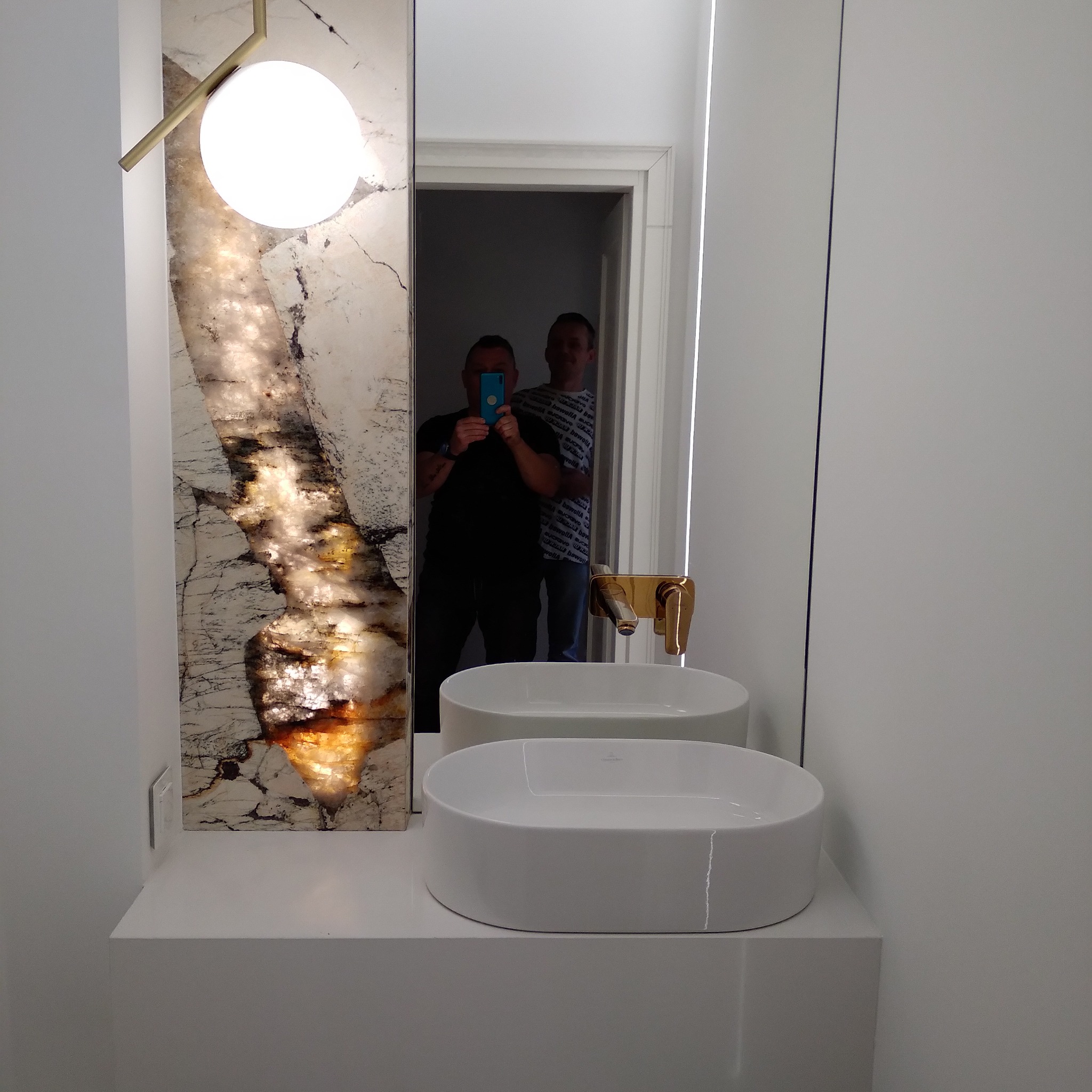 Nowoczesna łazienka z dwoma białymi umywalkami nablatowymi, złotą armaturą i podświetlanym panelem z imitacją kamienia naturalnego obok lustra, w którym odbijają się dwie osoby robiące zdjęcie.