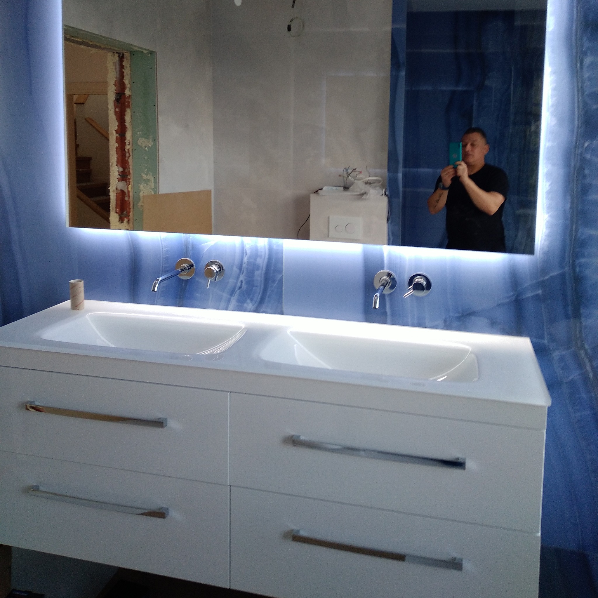 Nowoczesna łazienka z podwójną białą umywalką na szafce z czterema szufladami, chromowanymi uchwytami, podświetlanym lustrem i błękitną mozaiką na ścianie, widoczne fragmenty niedokończonego...