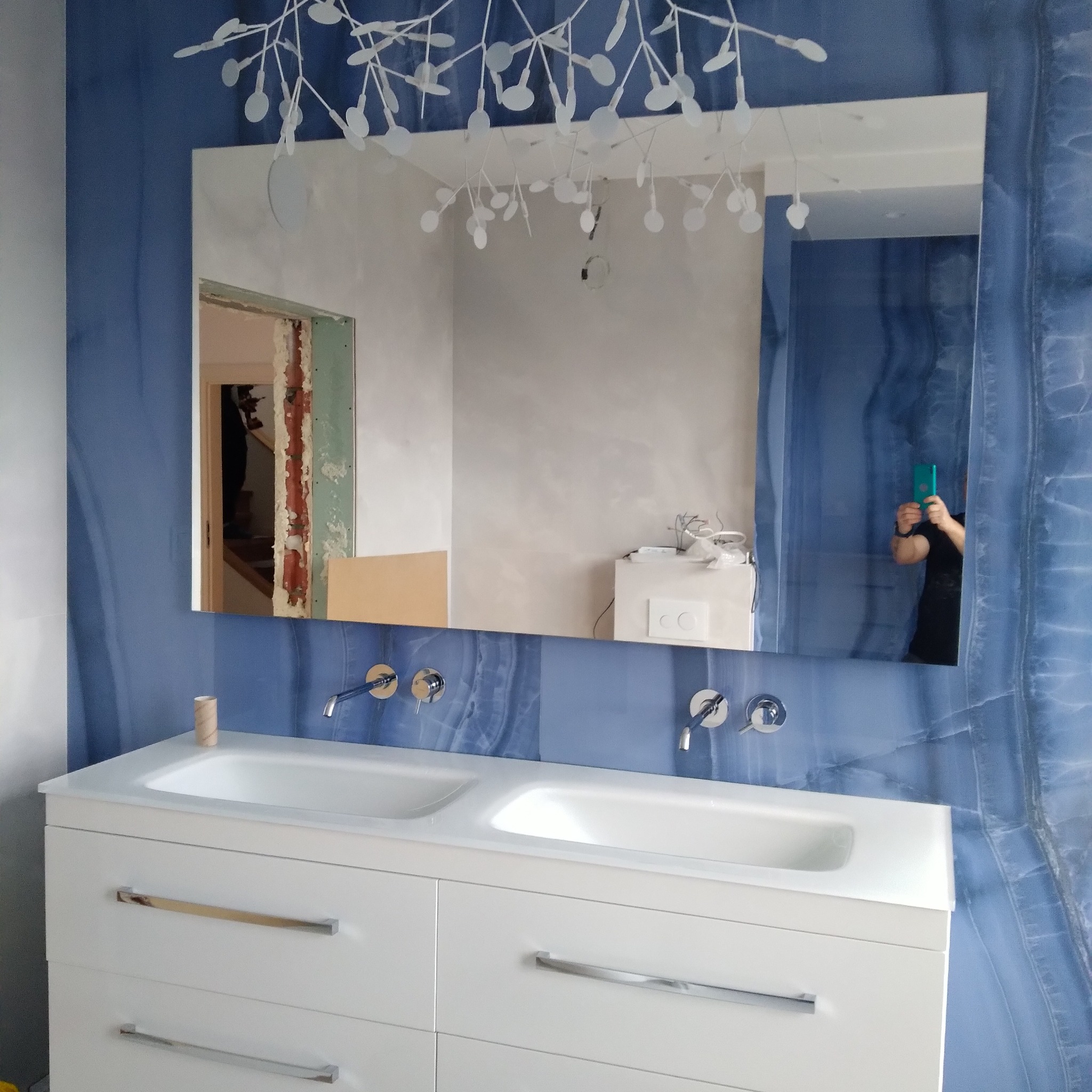 Nowoczesna łazienka z umywalką nablatową, bateriami ściennymi i lustrem, w którym odbija się osoba robiąca zdjęcie telefonem oraz widoczne prace remontowe w sąsiednim pomieszczeniu.