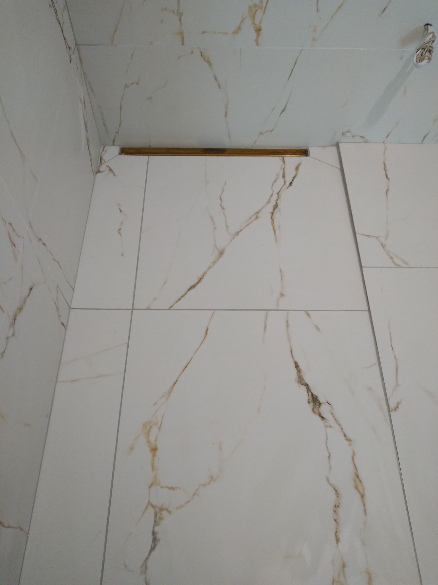 Białe płytki imitujące marmur z delikatnymi złotymi żyłkami, ułożone na ścianach i suficie łazienki, z widoczną złotą listwą dekoracyjną i wystającymi kablami oświetleniowymi.