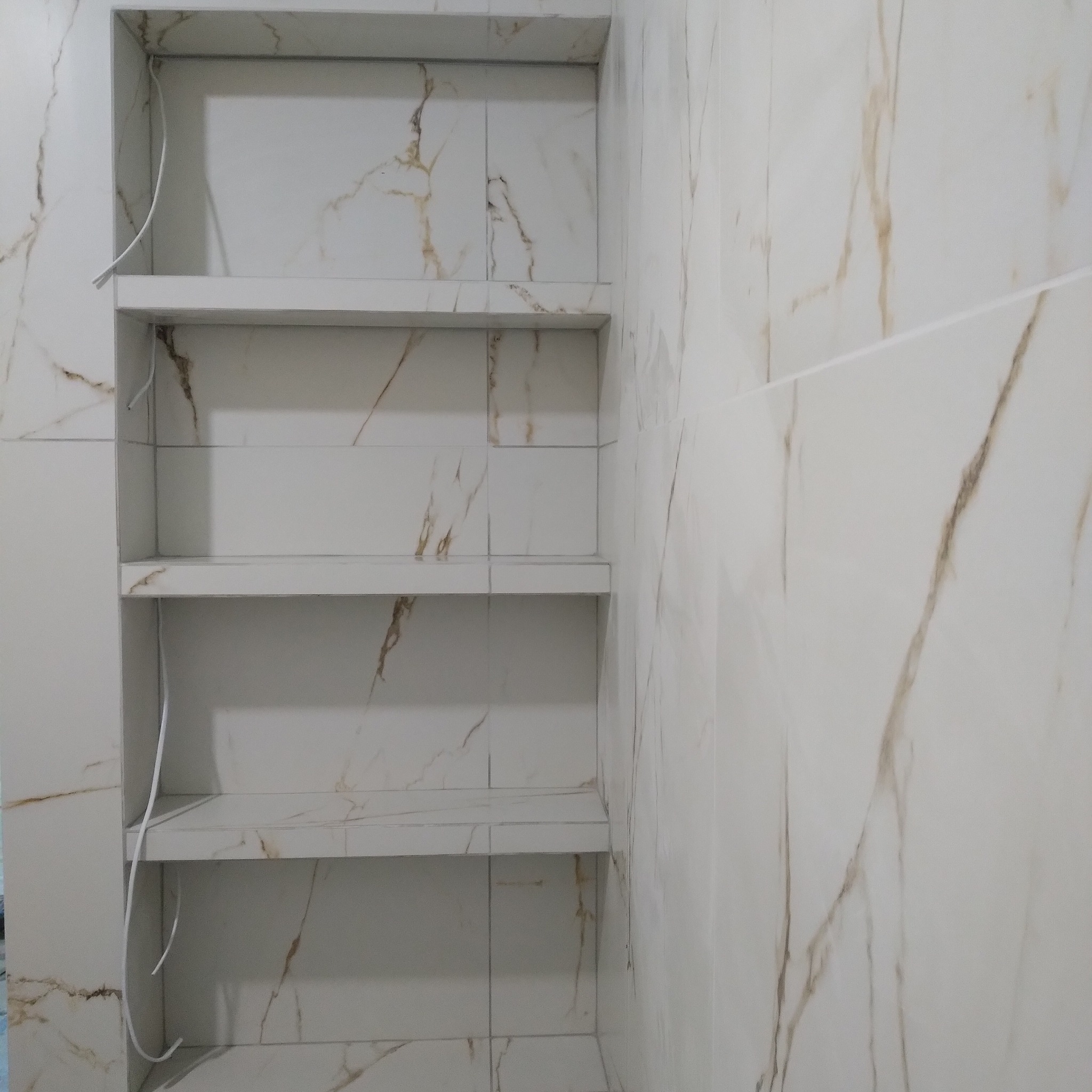 Wnęka z półkami wyłożona białymi płytkami imitującymi marmur, z widocznymi przewodami elektrycznymi, prawdopodobnie przygotowana pod oświetlenie.