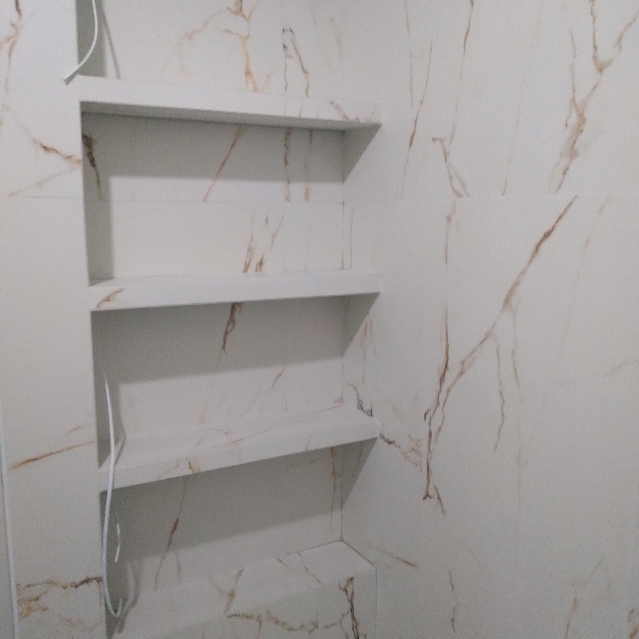 Wnęka z trzema półkami wyłożona płytkami imitującymi marmur, w łazience o jasnej kolorystyce.