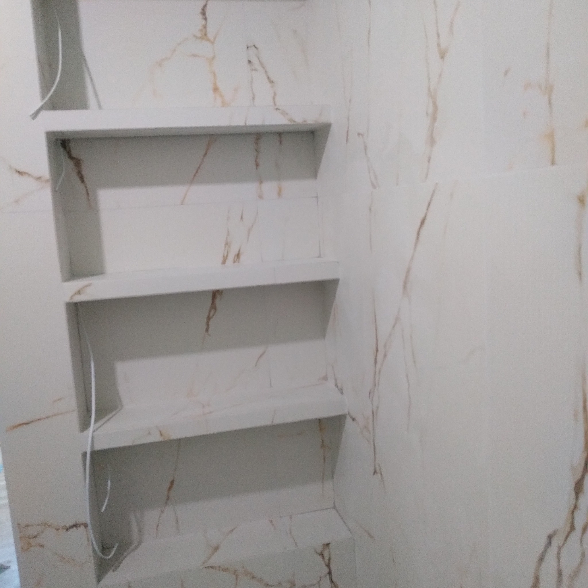 Wnęka z trzema półkami wyłożona płytkami imitującymi marmur w odcieniach bieli i beżu, z widocznymi kablami elektrycznymi.