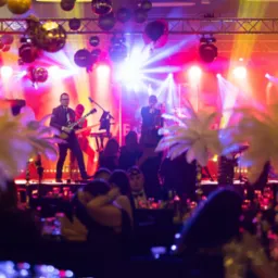 Scena z muzykami podczas występu na tle dekoracji z kulami dyskotekowymi, widziana zza stolików gości.