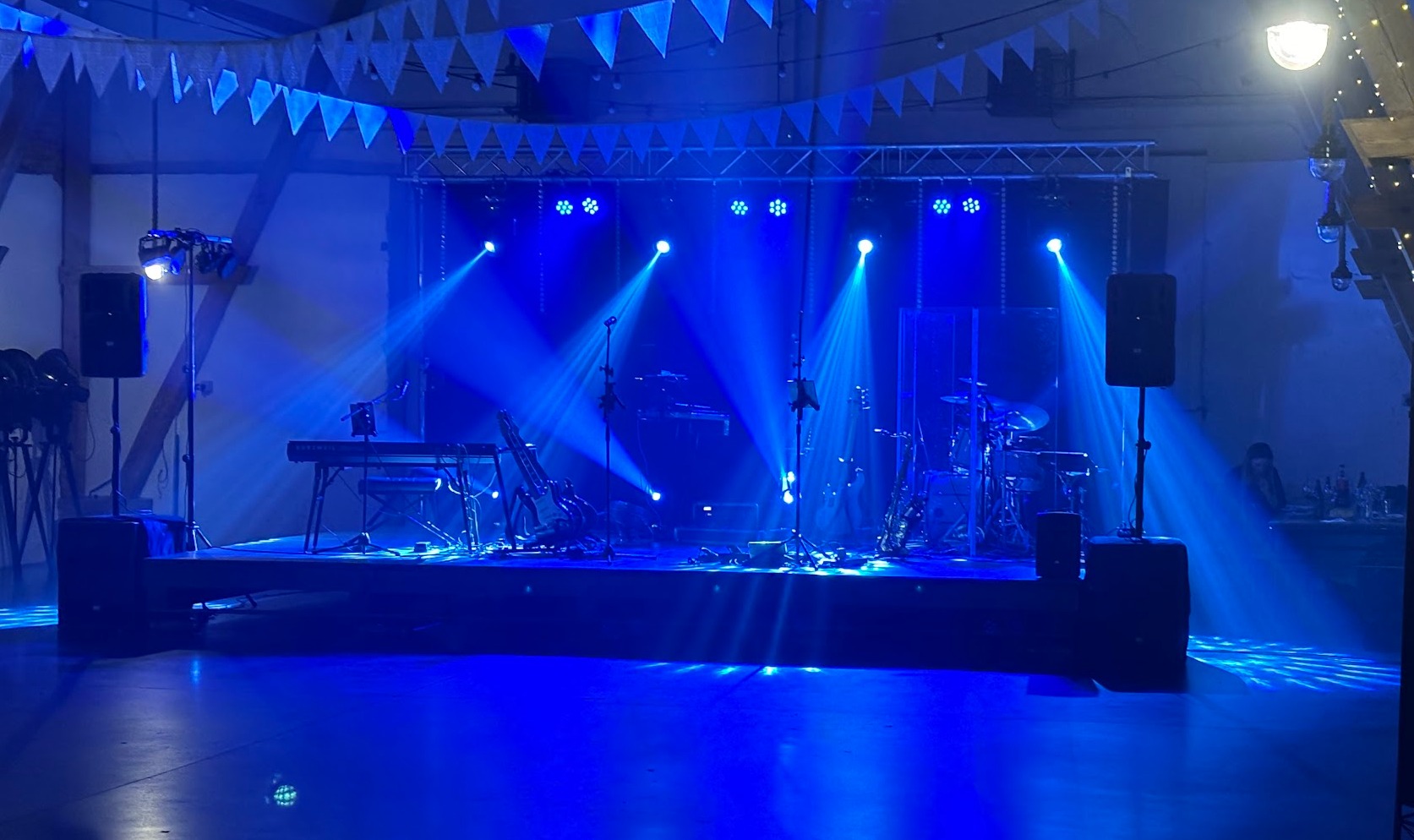 Scena oświetlona niebieskimi reflektorami, przygotowana do występu z instrumentami: keyboard, perkusja, gitara i saksofon.
