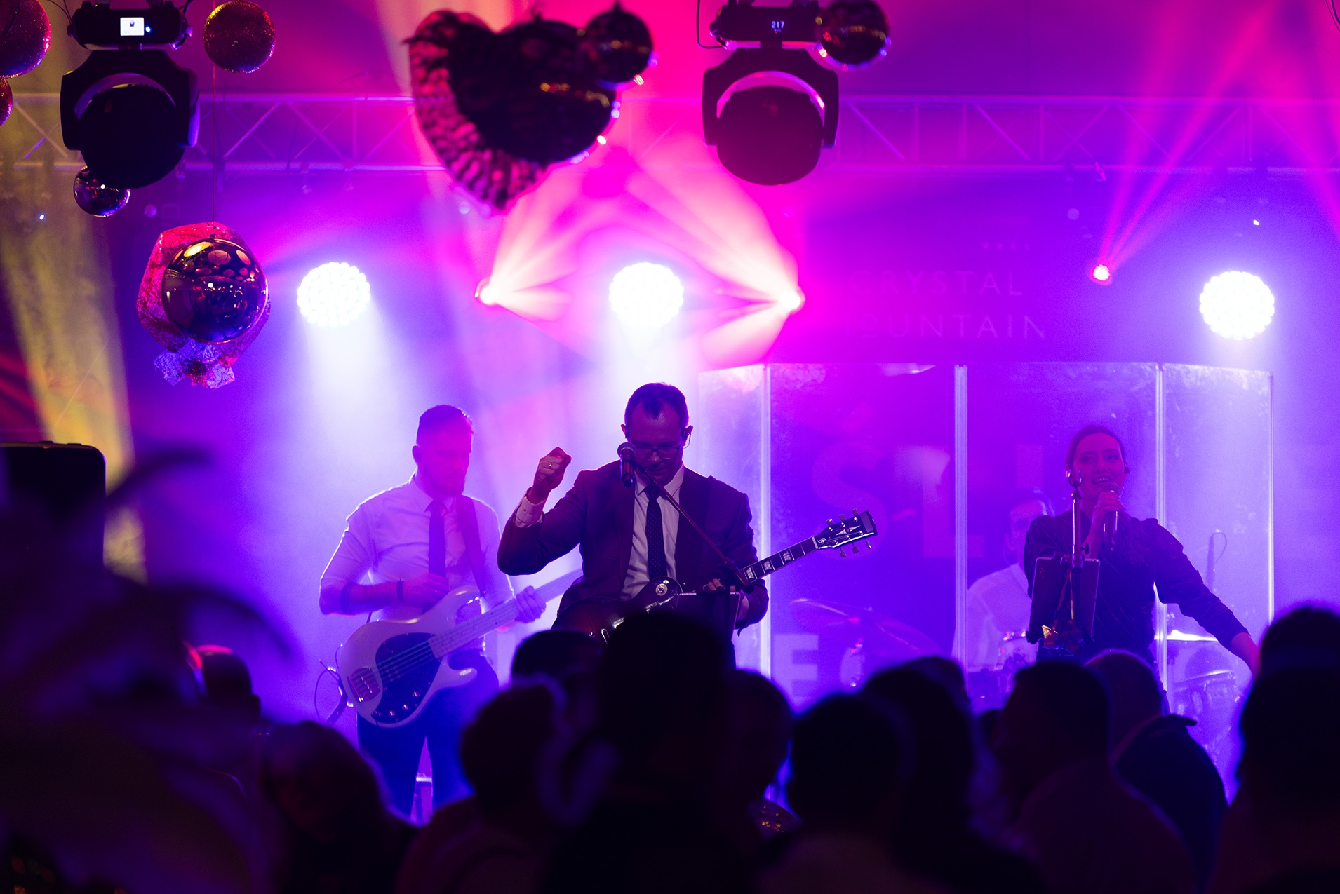 Zespół muzyczny grający koncert na scenie oświetlonej fioletowymi światłami, widoczni gitarzysta w garniturze i wokalistka, z przodu widownia.
