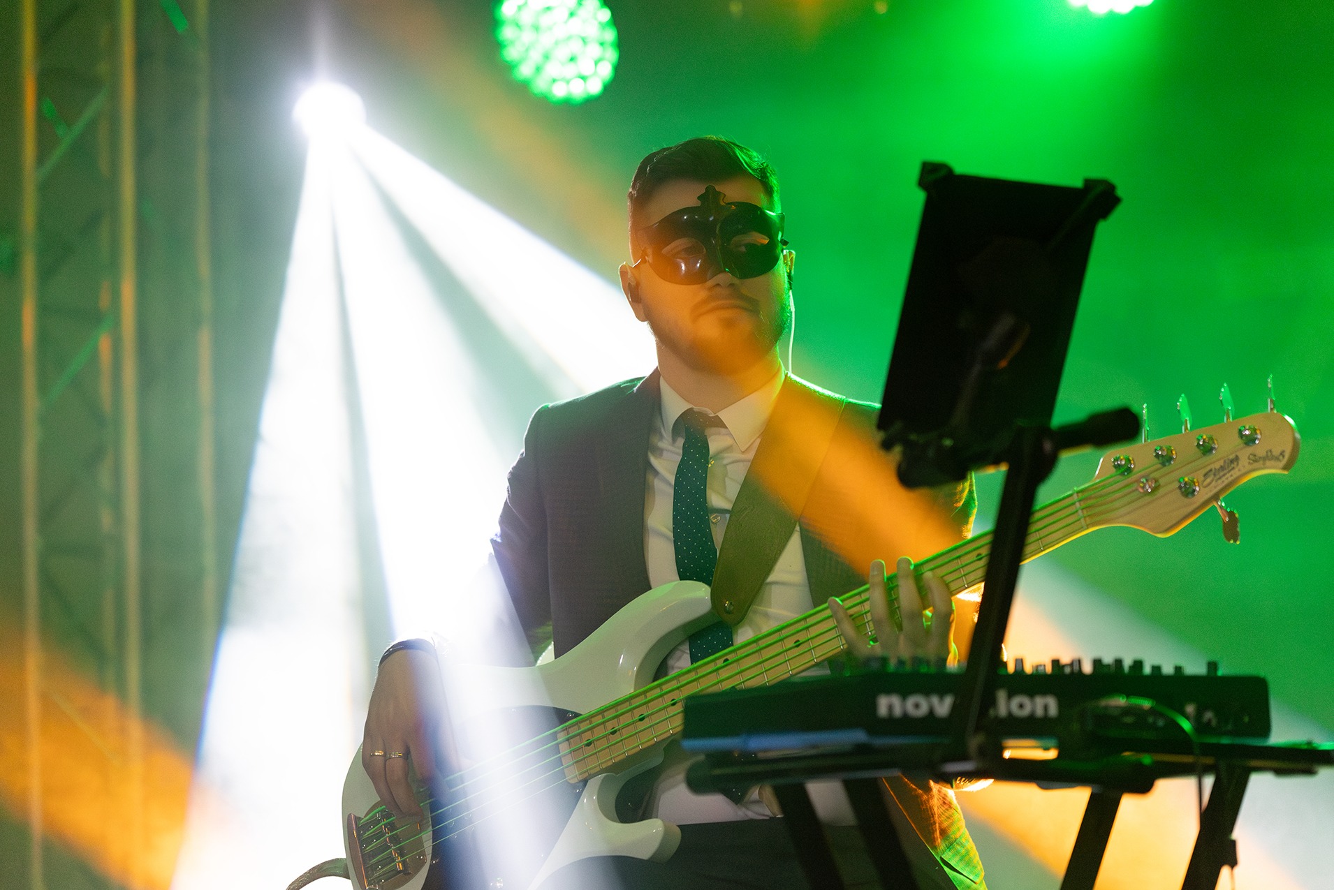 Basista w masce na scenie, grający na białym basie Sterling StingRay, oświetlony zielonym i białym światłem, z klawiszami Novation w tle.