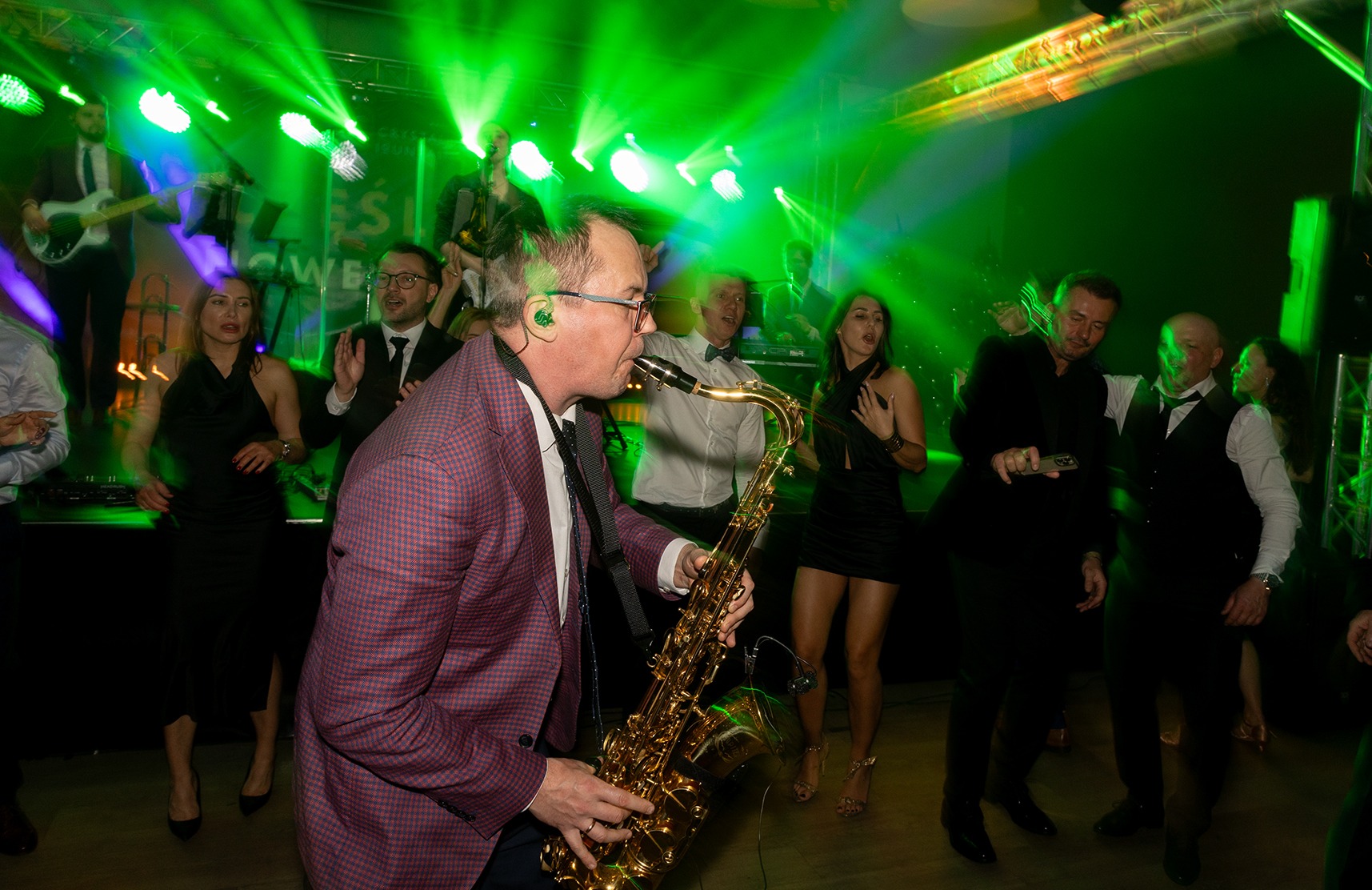 Saksofonista w bordowej marynarce w kratę gra na saksofonie na scenie oświetlonej zielonymi reflektorami, w tle wokalistki i muzycy.