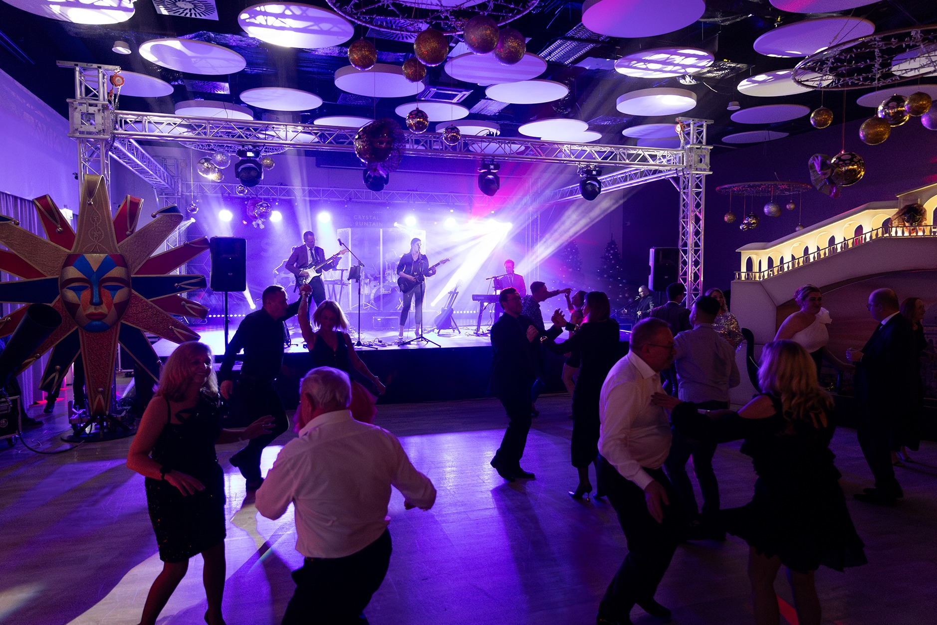 Zespół muzyczny na scenie oświetlonej fioletowym światłem, grający dla tańczących gości podczas imprezy okolicznościowej z dekoracjami sufitowymi i maską wenecką w tle.