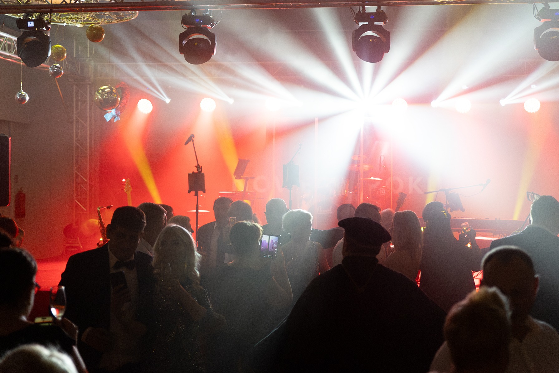 Widok na scenę z oświetleniem i sylwetki publiczności podczas występu, w tle widoczne instrumenty: saksofon, gitara basowa i keyboard.