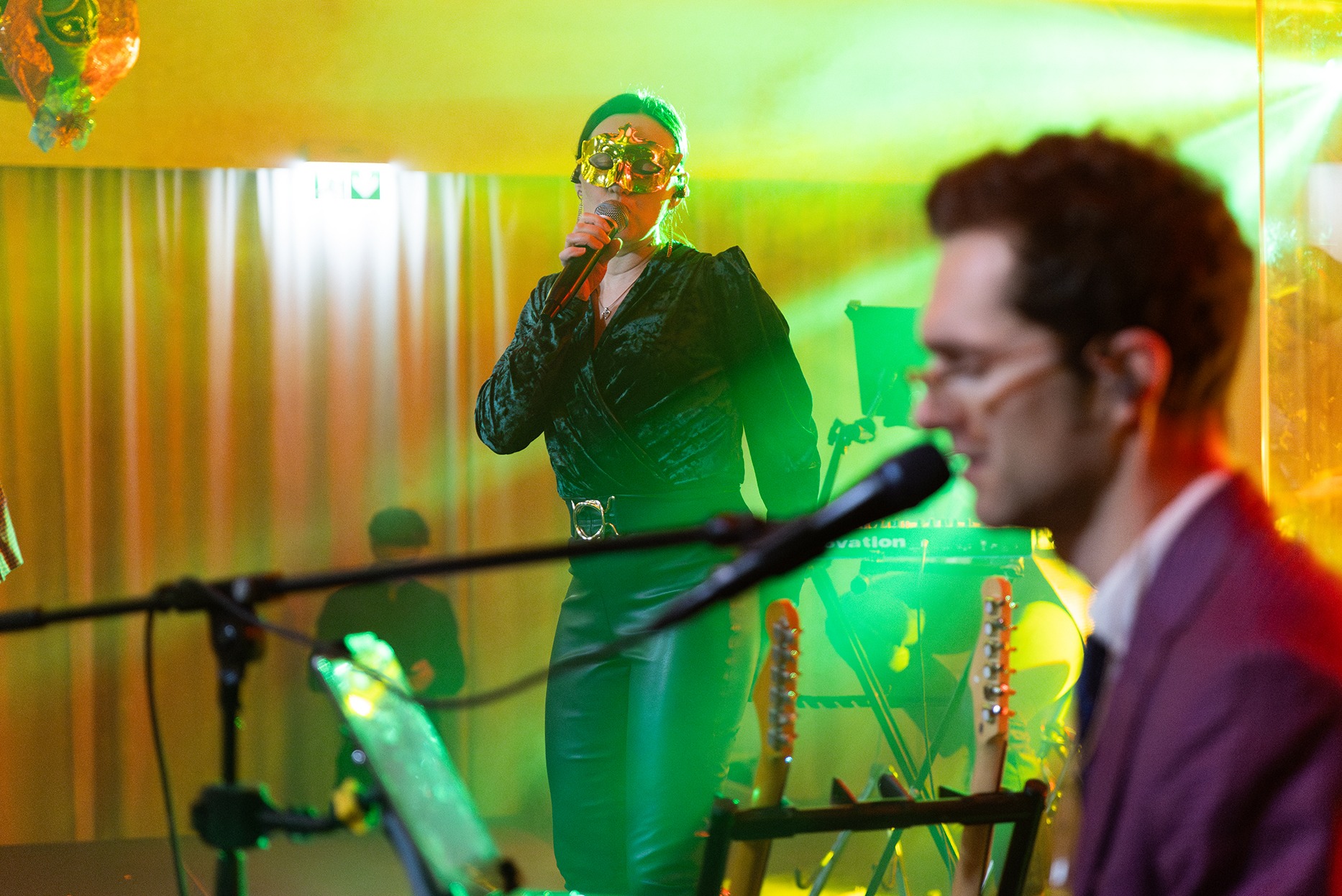 Wokalistka w złotej masce śpiewa do mikrofonu, obok muzyk z gitarą, oświetlenie sceniczne w odcieniach zieleni i żółci.