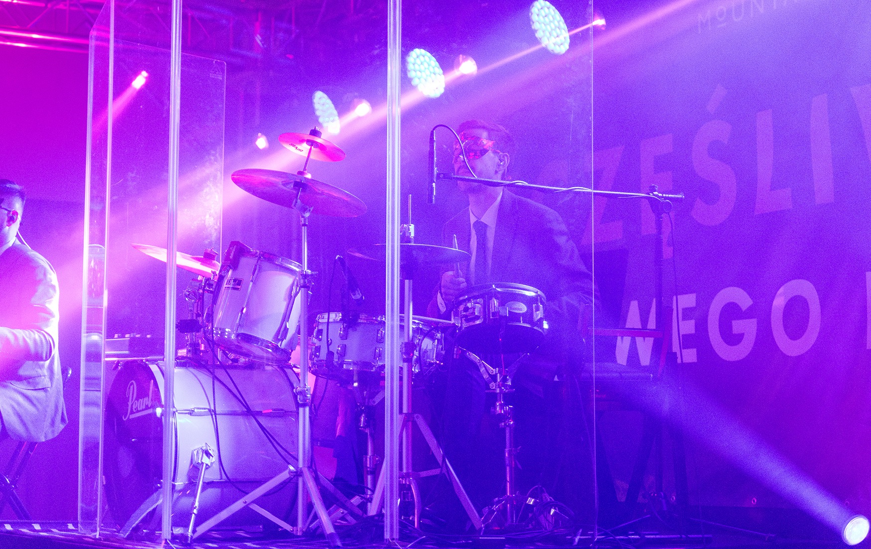 Perkusista w garniturze gra na perkusji Pearl za przezroczystą barierą, oświetlony fioletowymi światłami scenicznymi na tle z napisem.